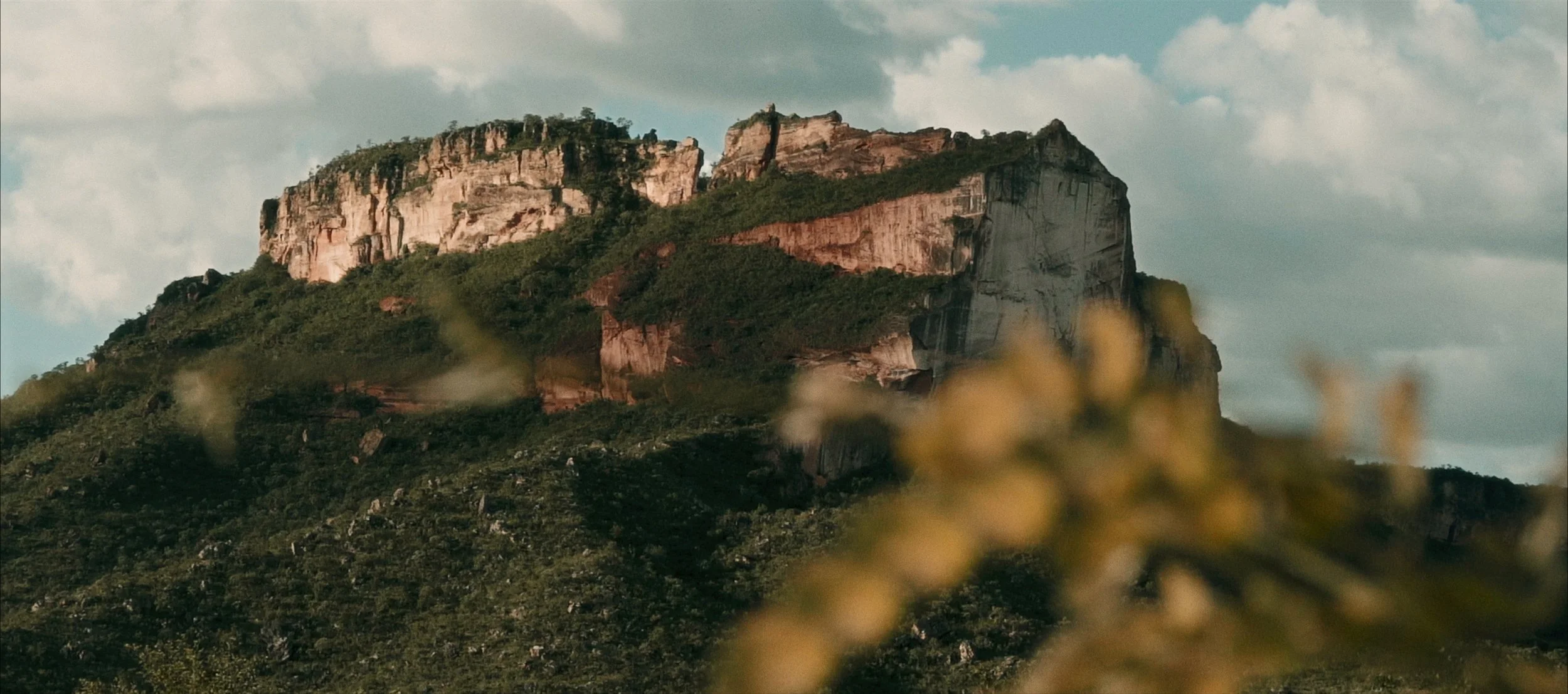 A rugged mountain with steep cliffs, covered in green vegetation, under a partly cloudy sky