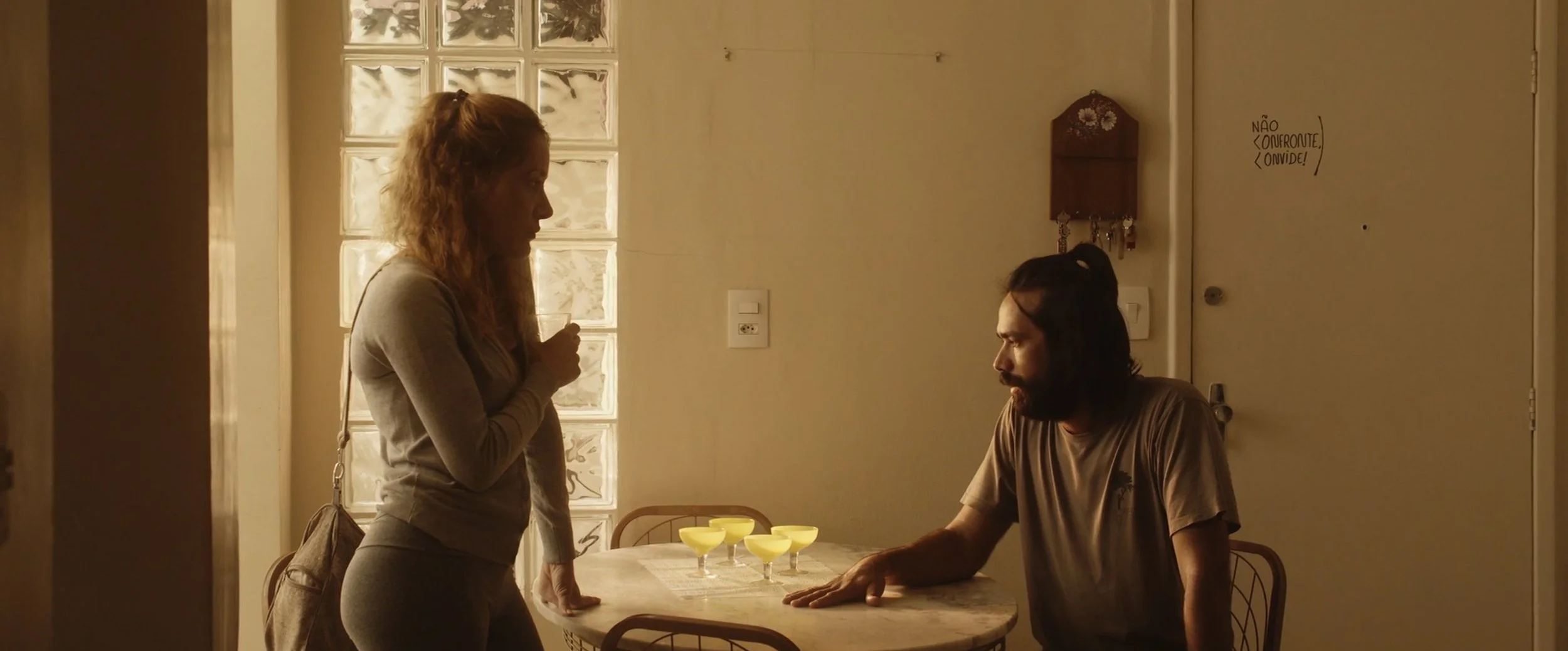 A woman standing and talking to a man sitting at a kitchen table with four yellow drinks, in a softly lit room with glass block window and a wall with small notes.