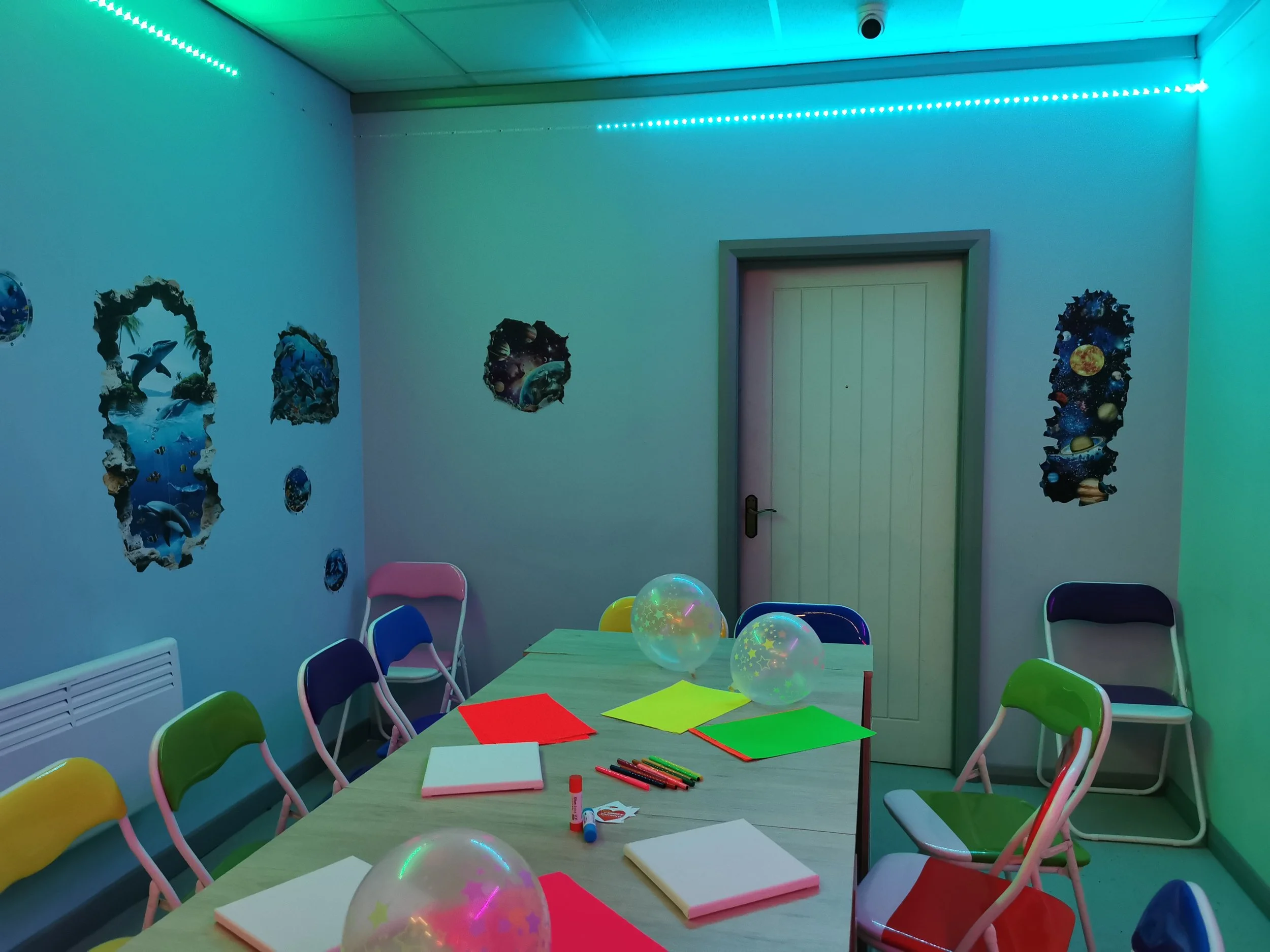 Colorful party or activity room with a wooden table, chairs, balloons, notebooks, and markers. The walls are decorated with  various themed posters and the room is lit with bluish-green LED lights.