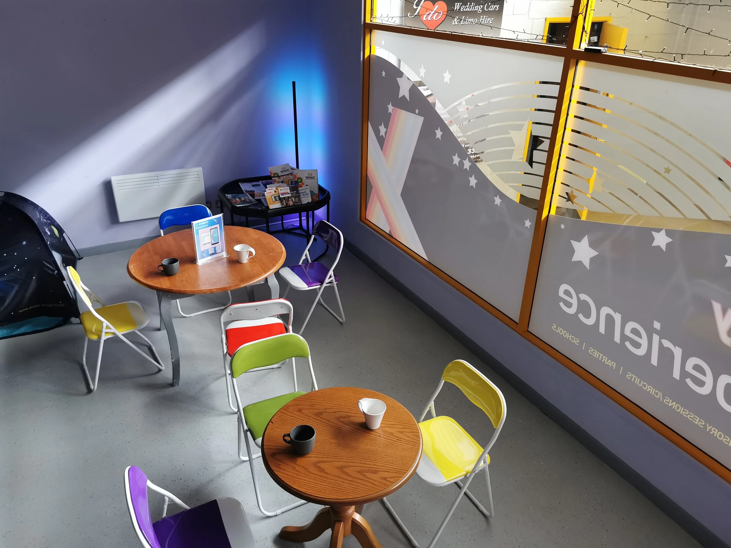 Interior of a coffee area with two wooden round tables, colorful chairs, and large window with store signage.