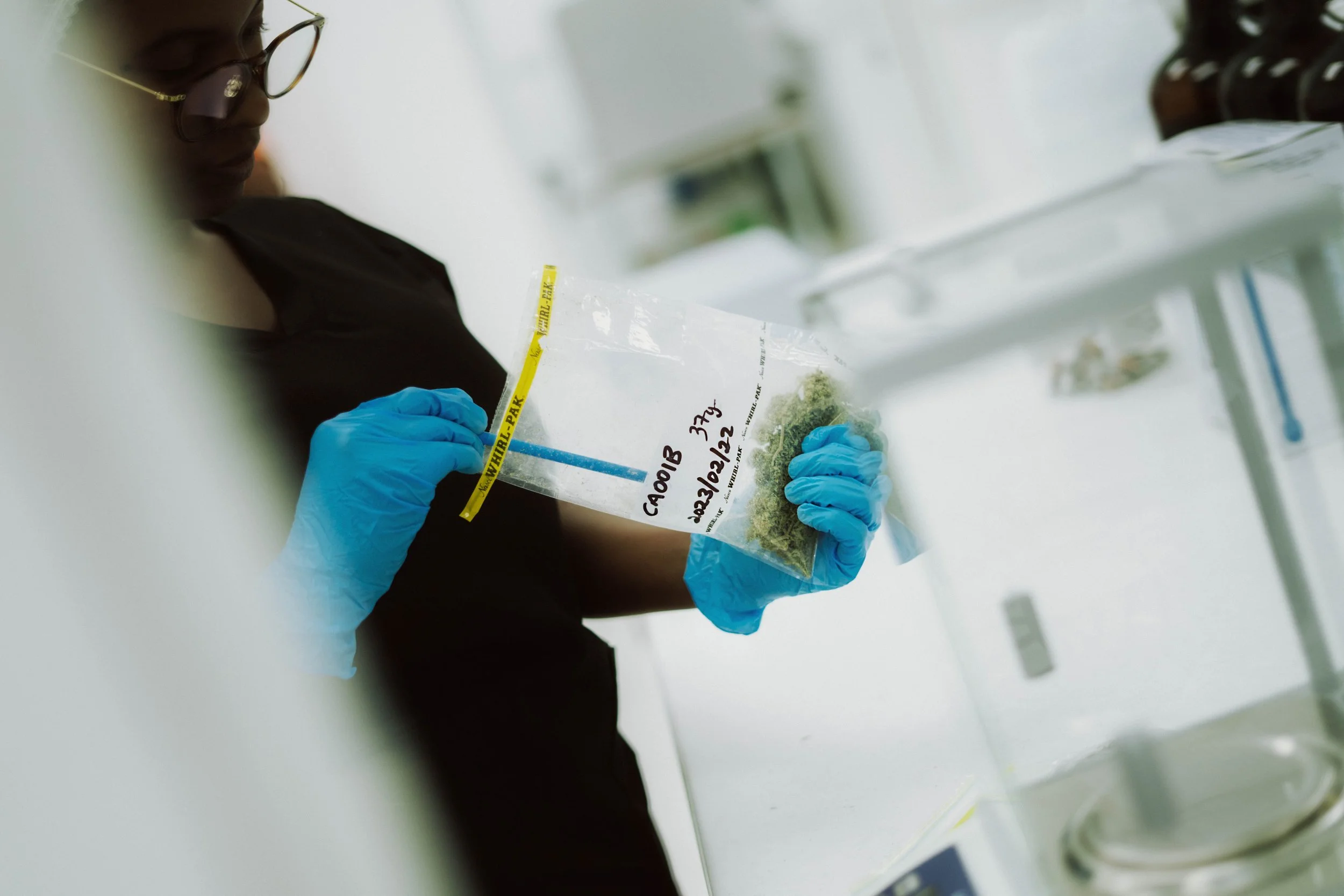 Person wearing blue gloves and glasses holding a transparent bag labeled with handwritten text in a laboratory setting.