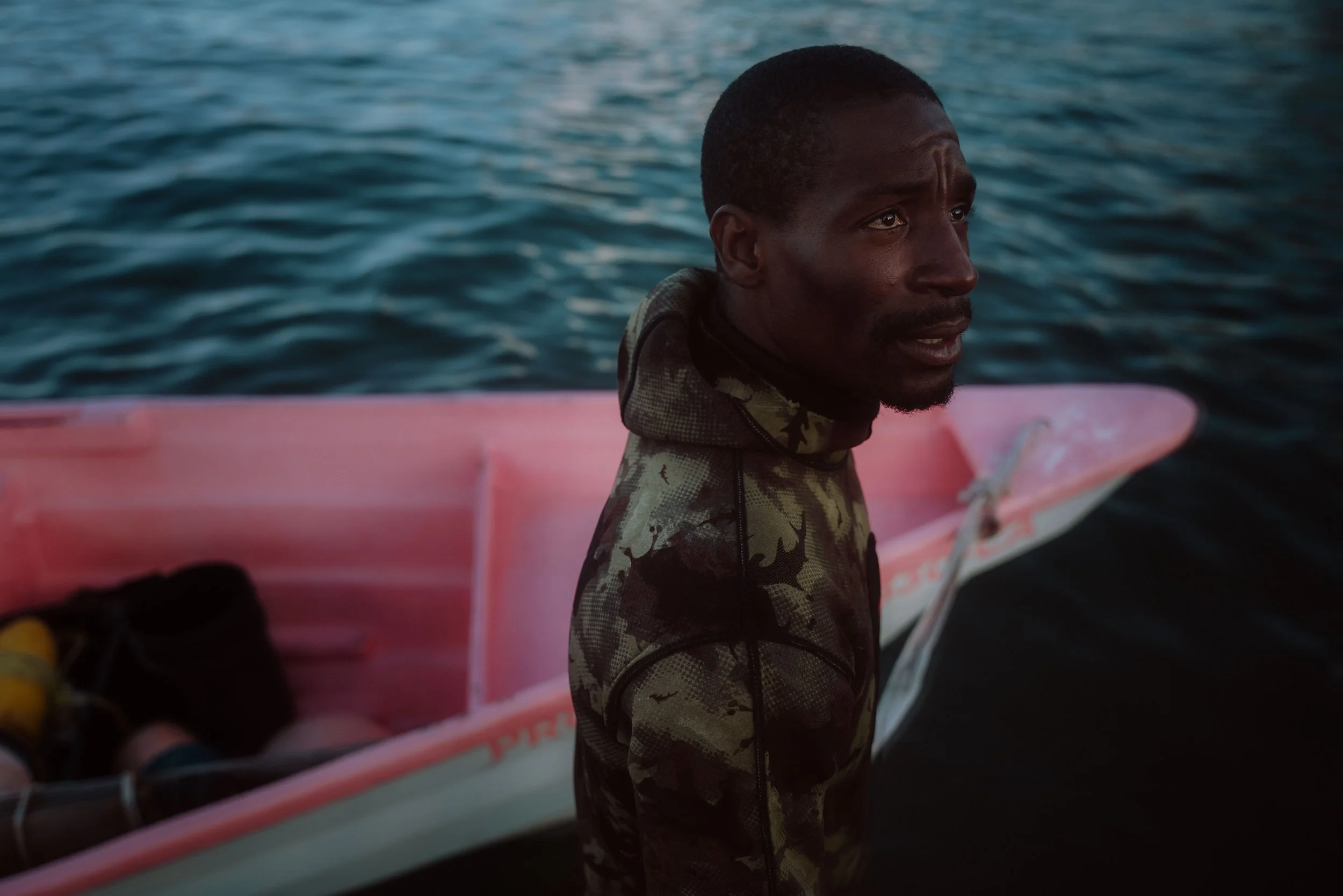 Man in a camouflage wetsuit standing beside a pink boat on the water.