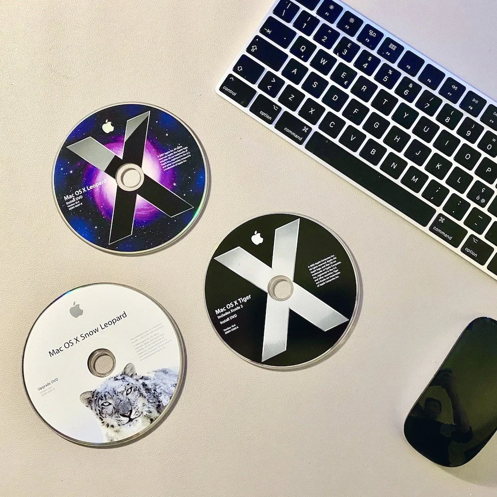 Tre dischi di installazione del sistema operativo Mac OS X Leopard, tra cui Snow Leopard, Lion e Tiger, posizionati su una scrivania accanto a una tastiera e un dispositivo nero, forse uno smartphone.