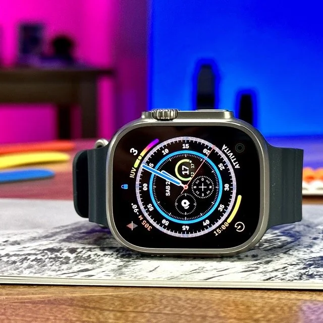 Orologio smart watch con quadrante digitale, posizionato su un foglio sul tavolo, con sfondo colorato di luci rosa e blu.