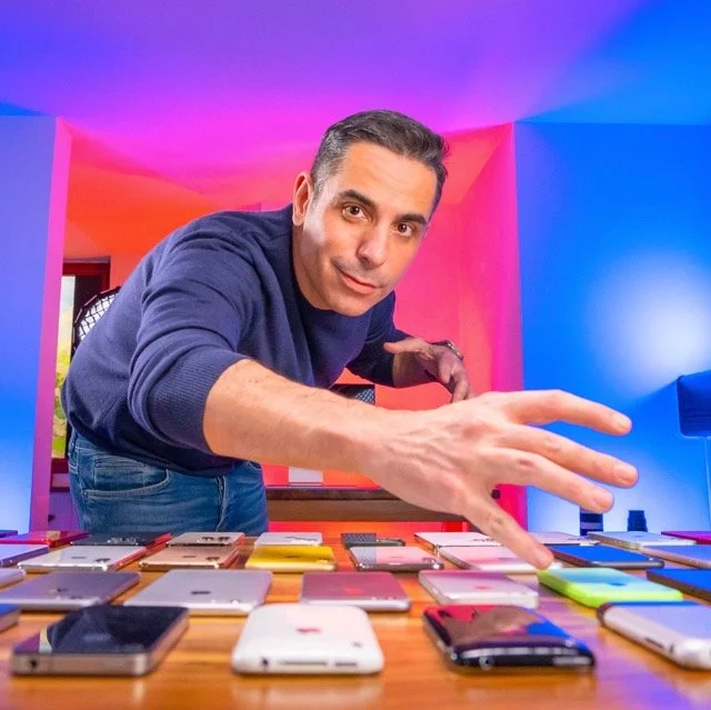 Uomo che sceglie tra vari smartphone disposti su un tavolo, in una stanza con pareti colorate fluorescenti che creano un'atmosfera vivace.