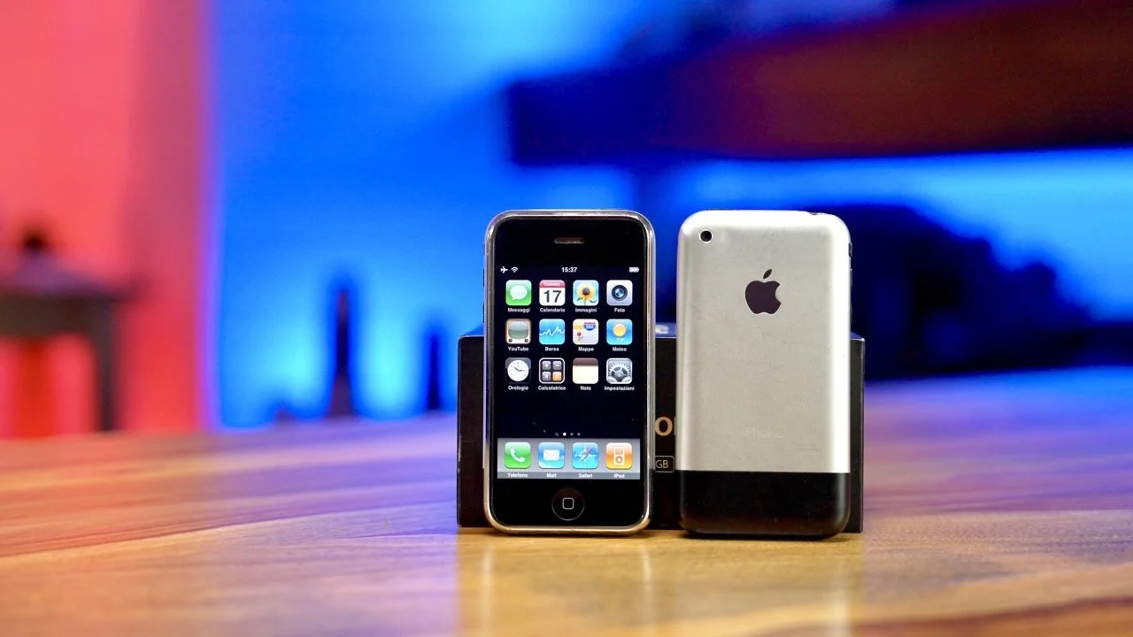 Un iPhone nero con schermo acceso e un iPhone bianco con retro, entrambi appoggiati a un supporto, su un tavolo di legno con sfondo illuminato di luci blu e rosse.