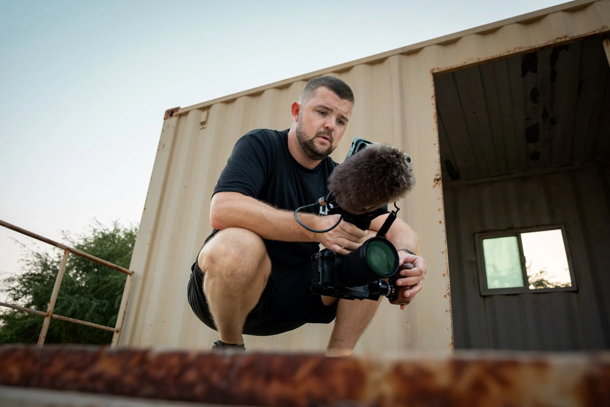 A man holding a camera rig stands outdoors near a modern building with glass windows and colorful blue and green vertical panels.