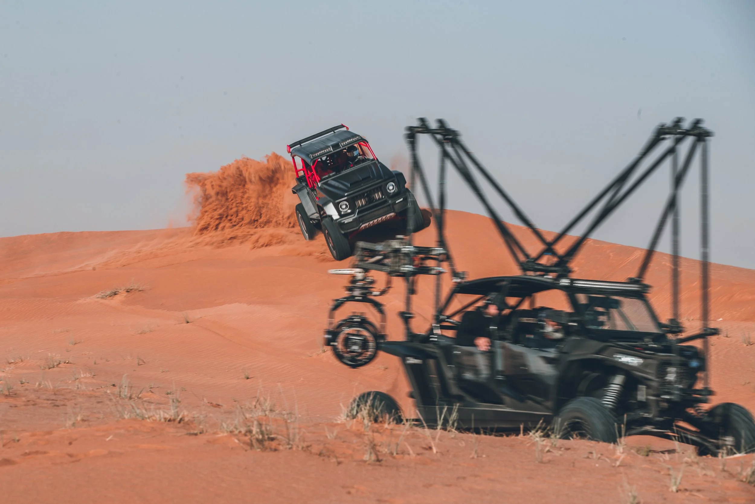 A black off-road vehicle is flying through the air as it climbs a sand dune in a desert, with loose sand trailing behind it.