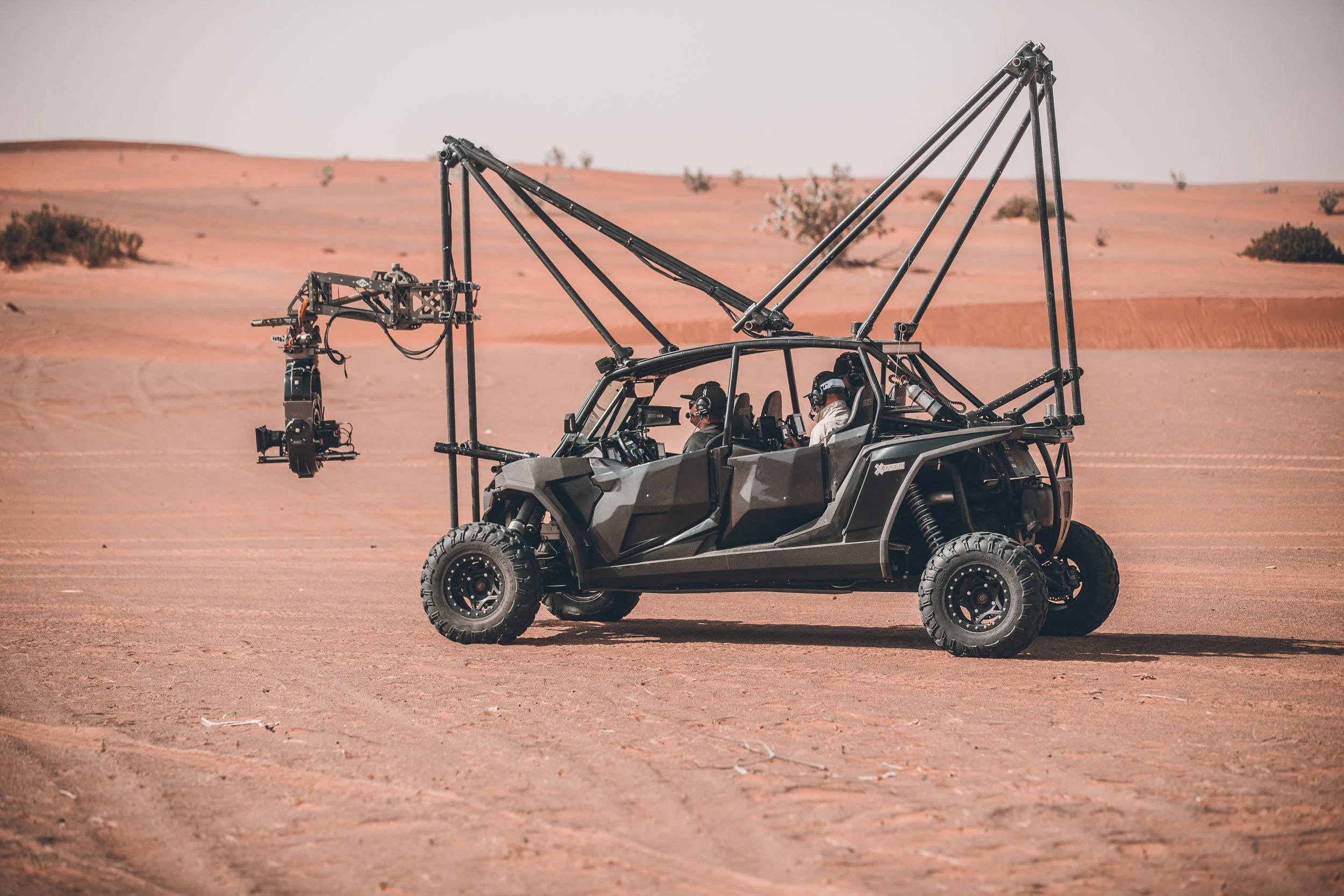 A black off-road vehicle with a mounted camera crane is in a desert landscape with sand dunes and sparse vegetation.
