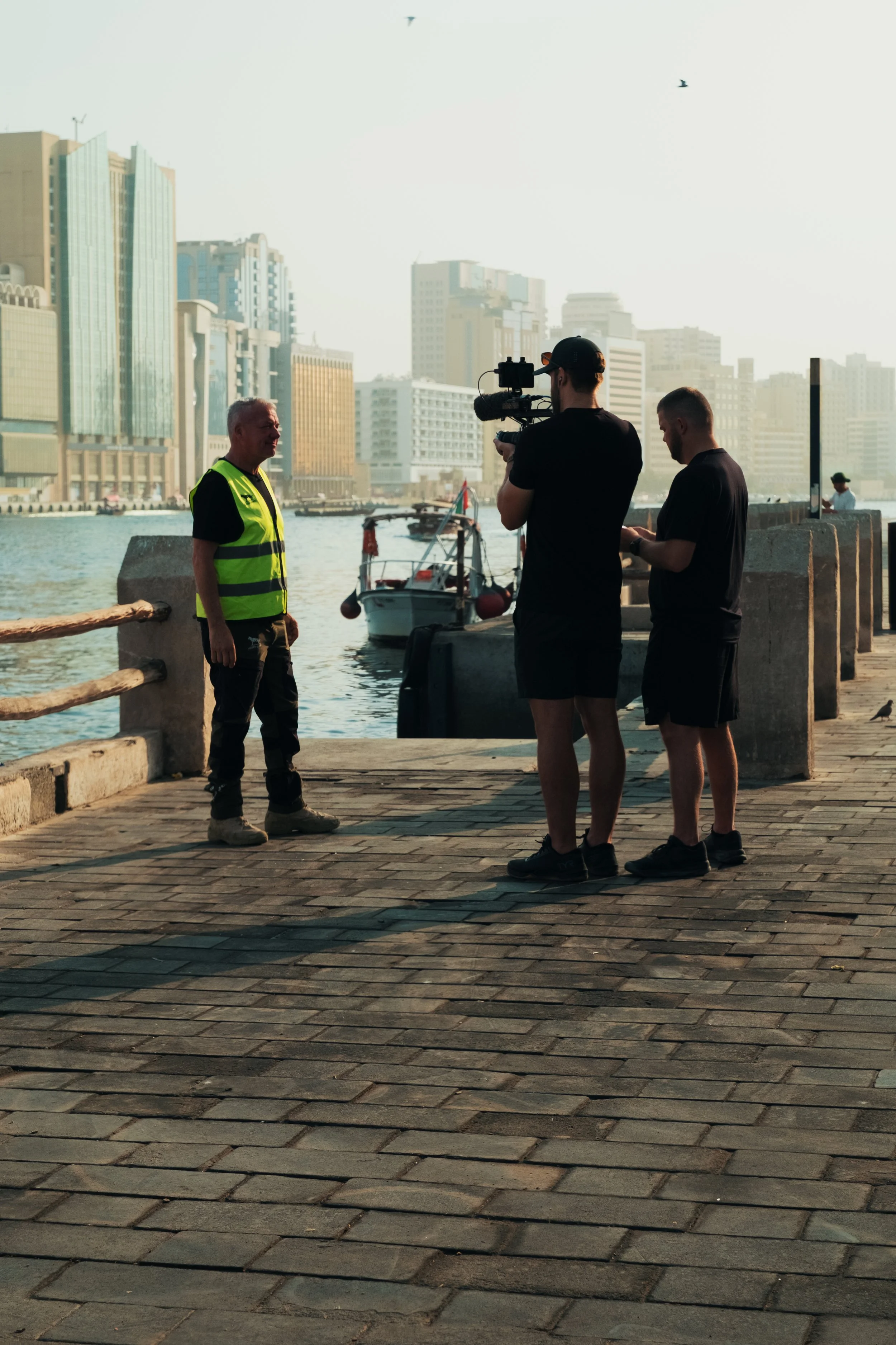 Three people filming a man in a high-visibility vest by the waterfront in a city, with tall modern buildings and boats in the background.