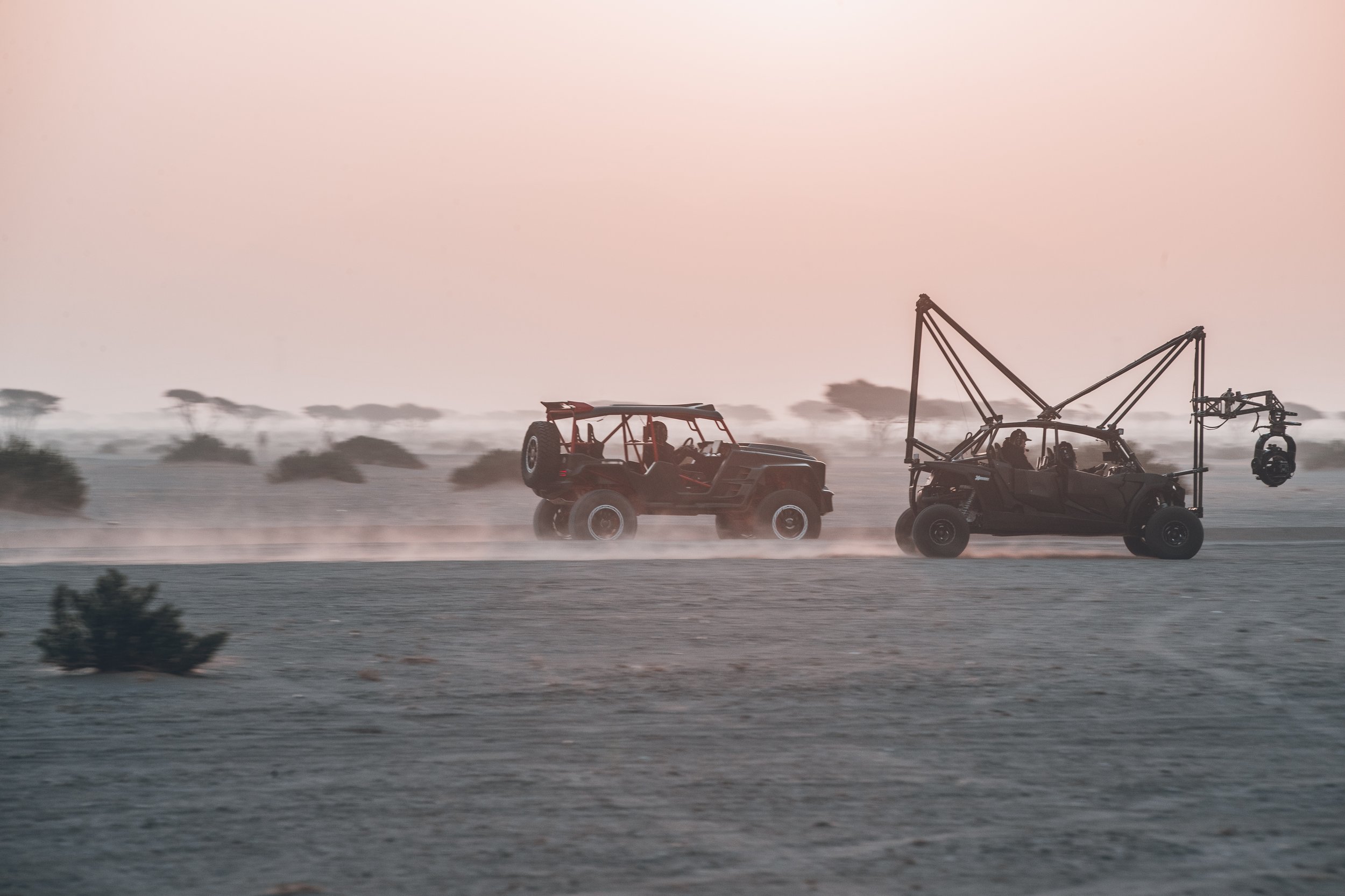 Two off-road vehicles, one with a camera rig, driving across a dusty desert landscape at sunset.