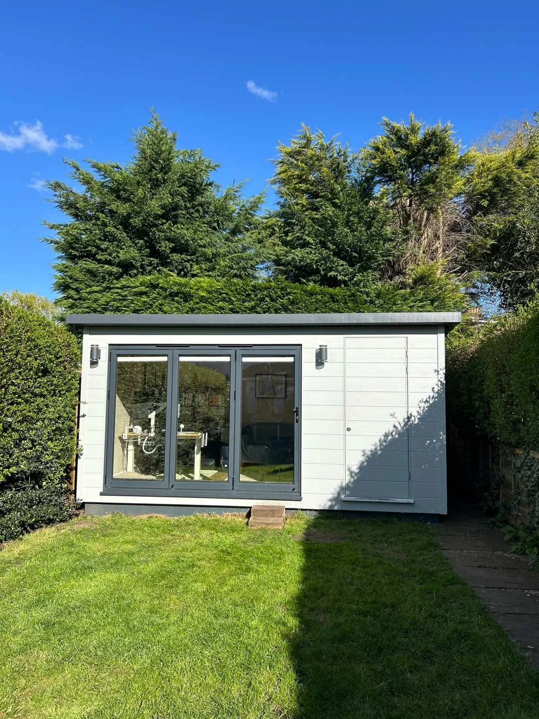 Front view of bespoke white composite-clad garden room with bi-fold doors, set within a green lawn and hedging