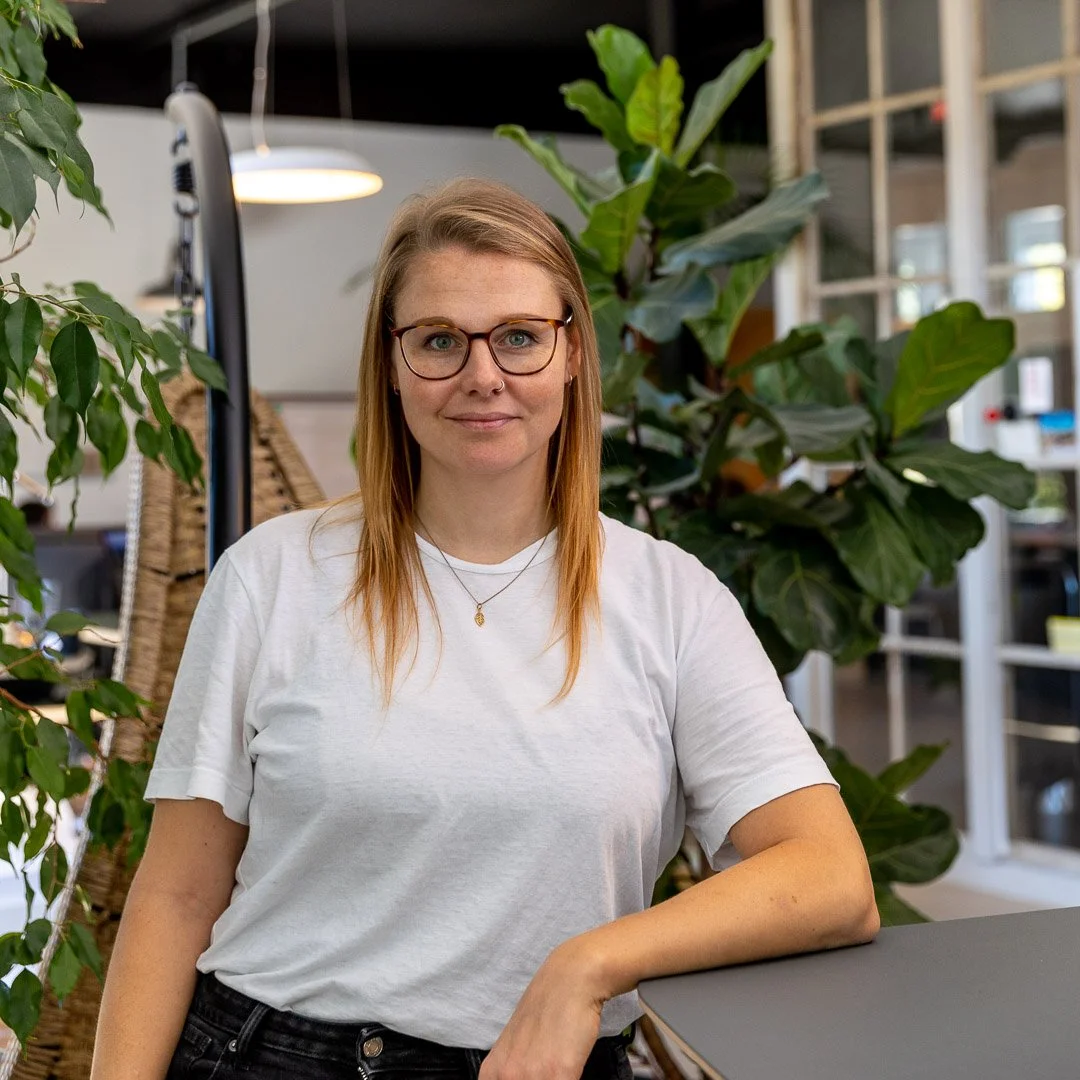 Junge Frau mit Brille und weissem T-Shirt sitzt in einem Raum mit großen Pflanzen im Hintergrund. Sie hat kupferrote Haare und trägt eine Halskette.