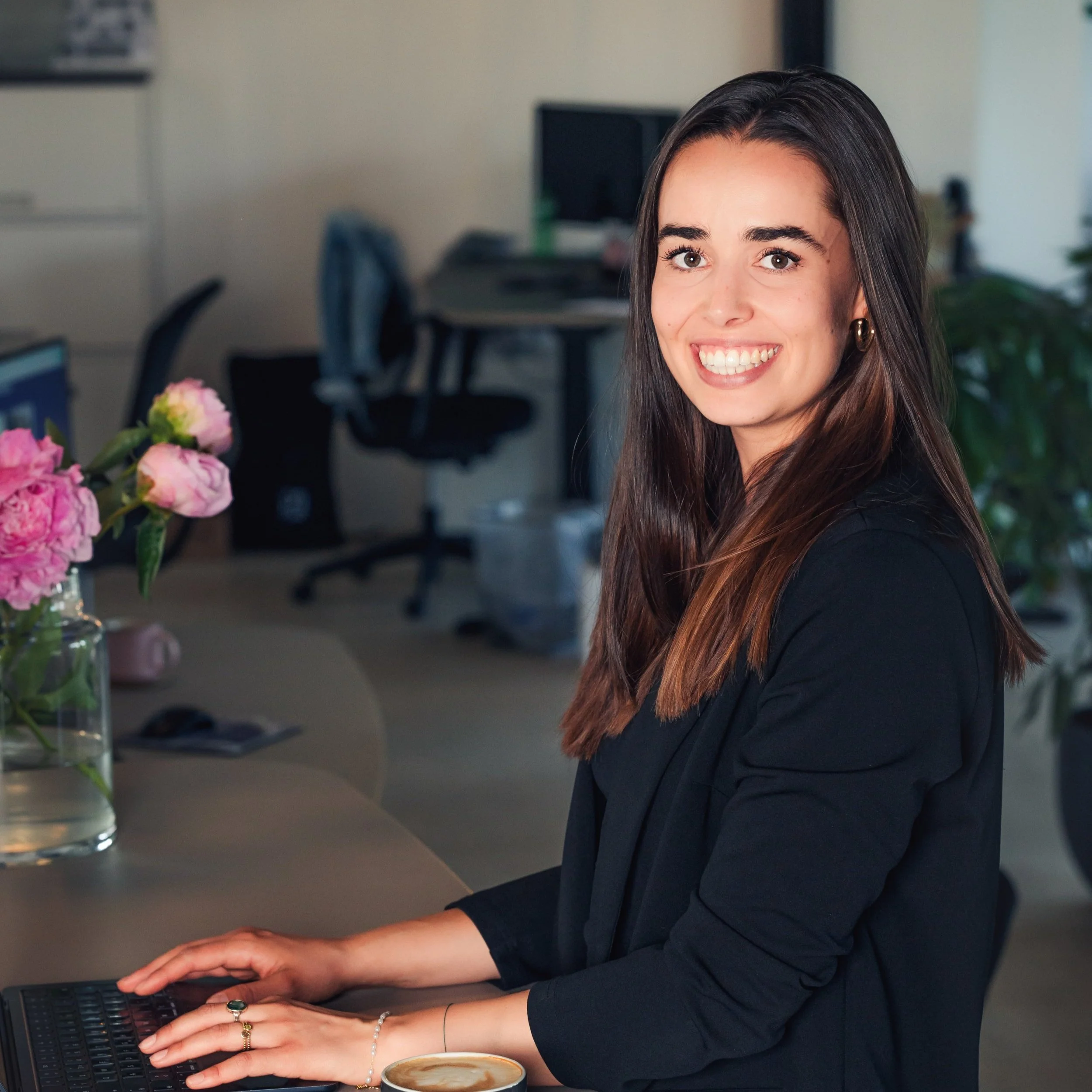 Eine lachende Frau arbeitet an einem Laptop in einem Büro, neben ihr eine Vase mit pinken Blumen.