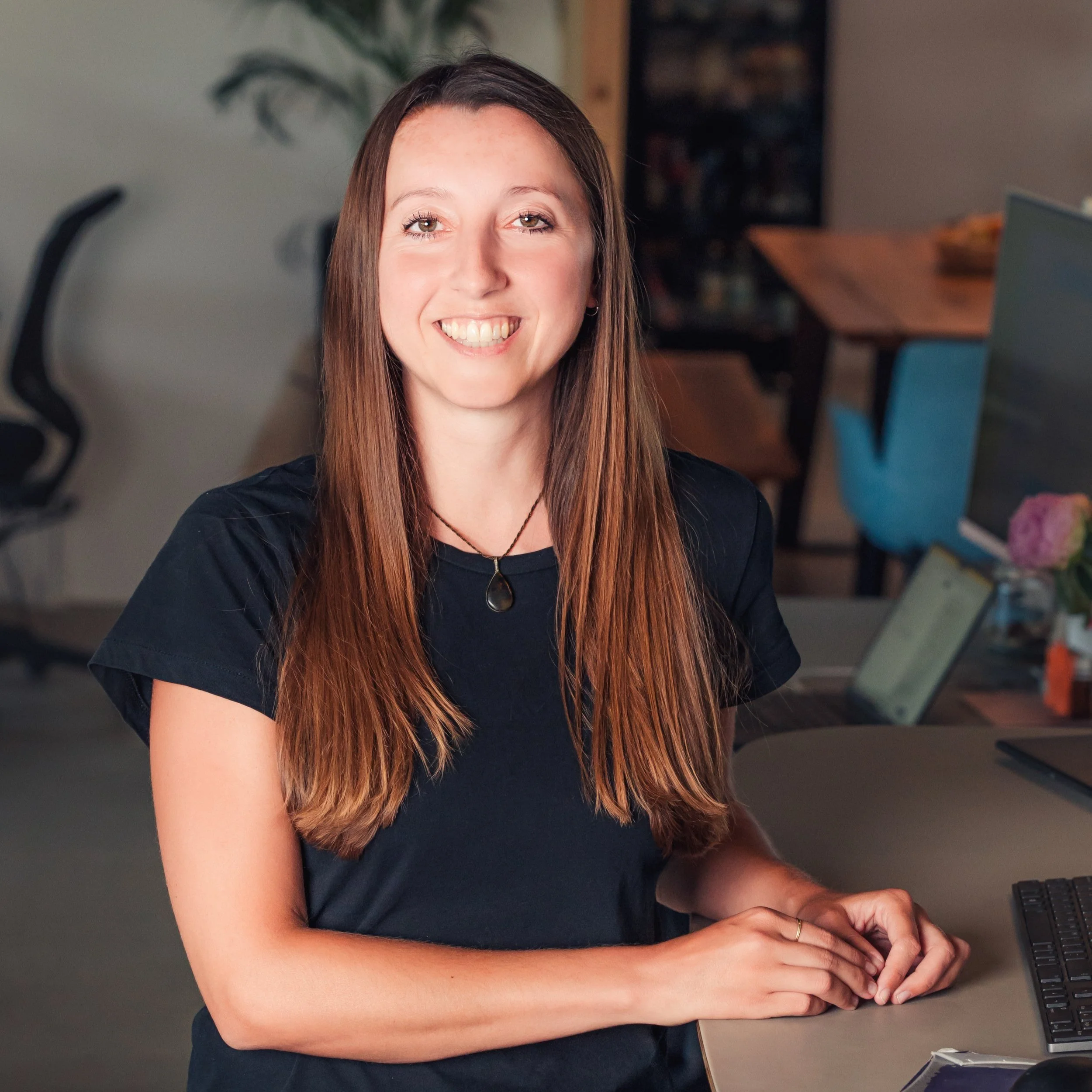 Lächelnde junge Frau mit langem braunem Haar, trägt ein schwarzes T-Shirt und eine Halskette, sitzt an einem Schreibtisch in einem modernen Büro.
