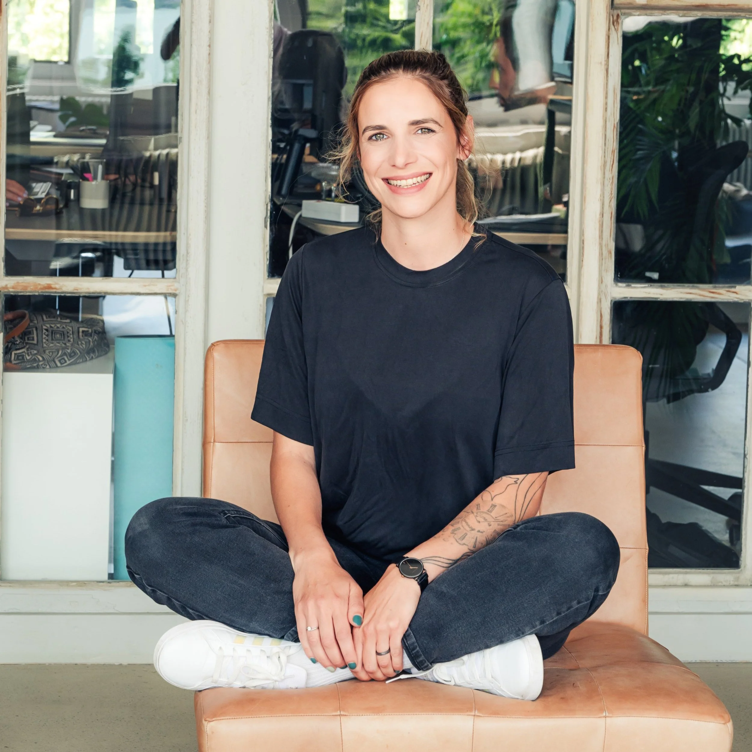 Junge Frau sitzt lachend auf einem Ledersofa, trägt ein schwarzes T-Shirt, schwarze Jeans, weiße Turnschuhe, eine Armbanduhr und hat Tattoos am linken Arm.