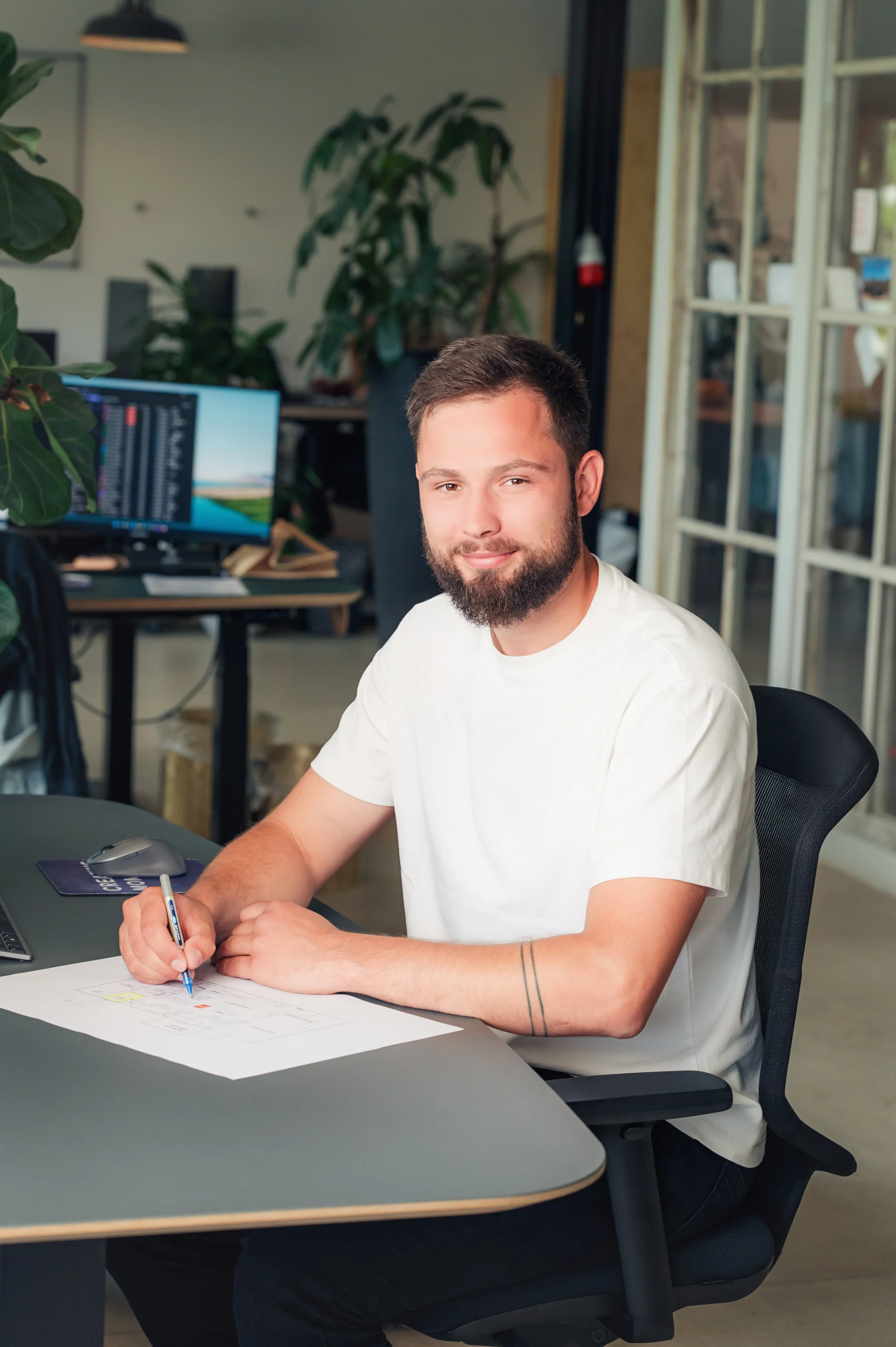 Ein junger Mann mit Bart und kurzen Haaren sitzt an einem Schreibtisch in einem modernen Büro, schreibt auf Papier mit einem blauen Stift, es gibt einen Computer, eine Maus und einen Bildschirm im Hintergrund.