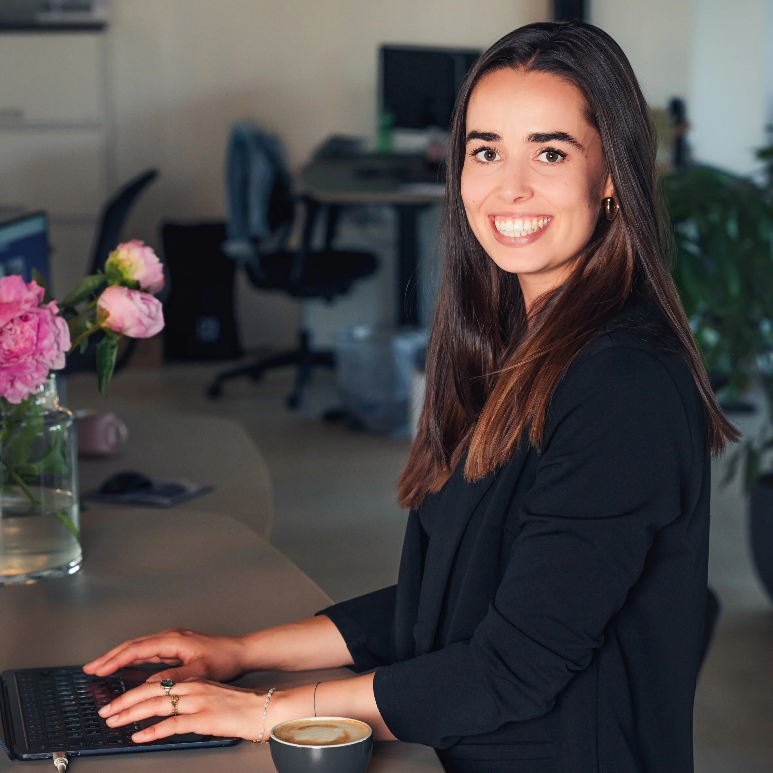 Eine lächelnde Frau sitzt an einem Tisch mit einem Laptop und einer Tasse Kaffee in einem Büro, neben einem Blumenvasen mit pinken Blumen.
