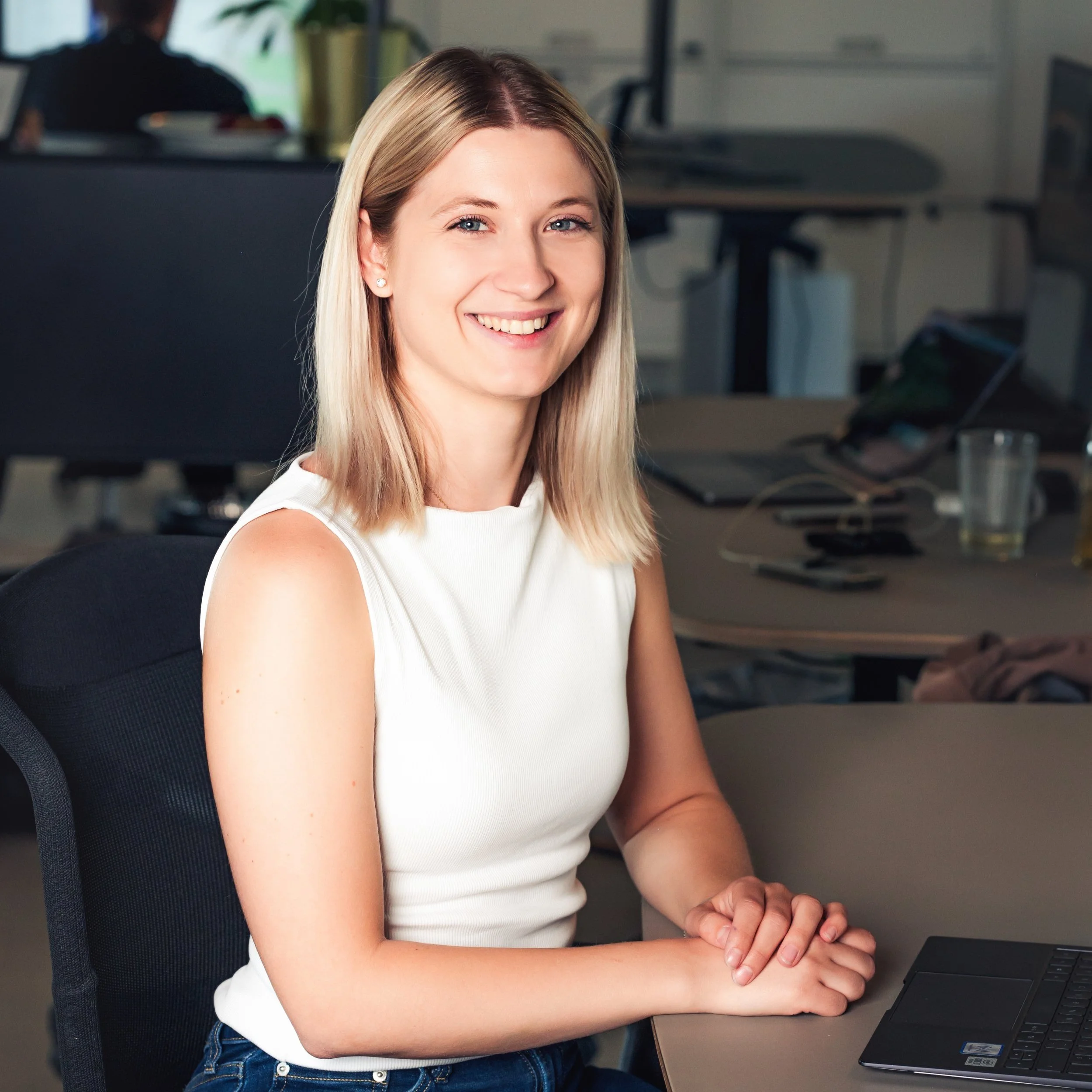Lächelnde Frau in einem Büro, sitzend an einem Tisch mit Laptop und Notizgeräten im Hintergrund.