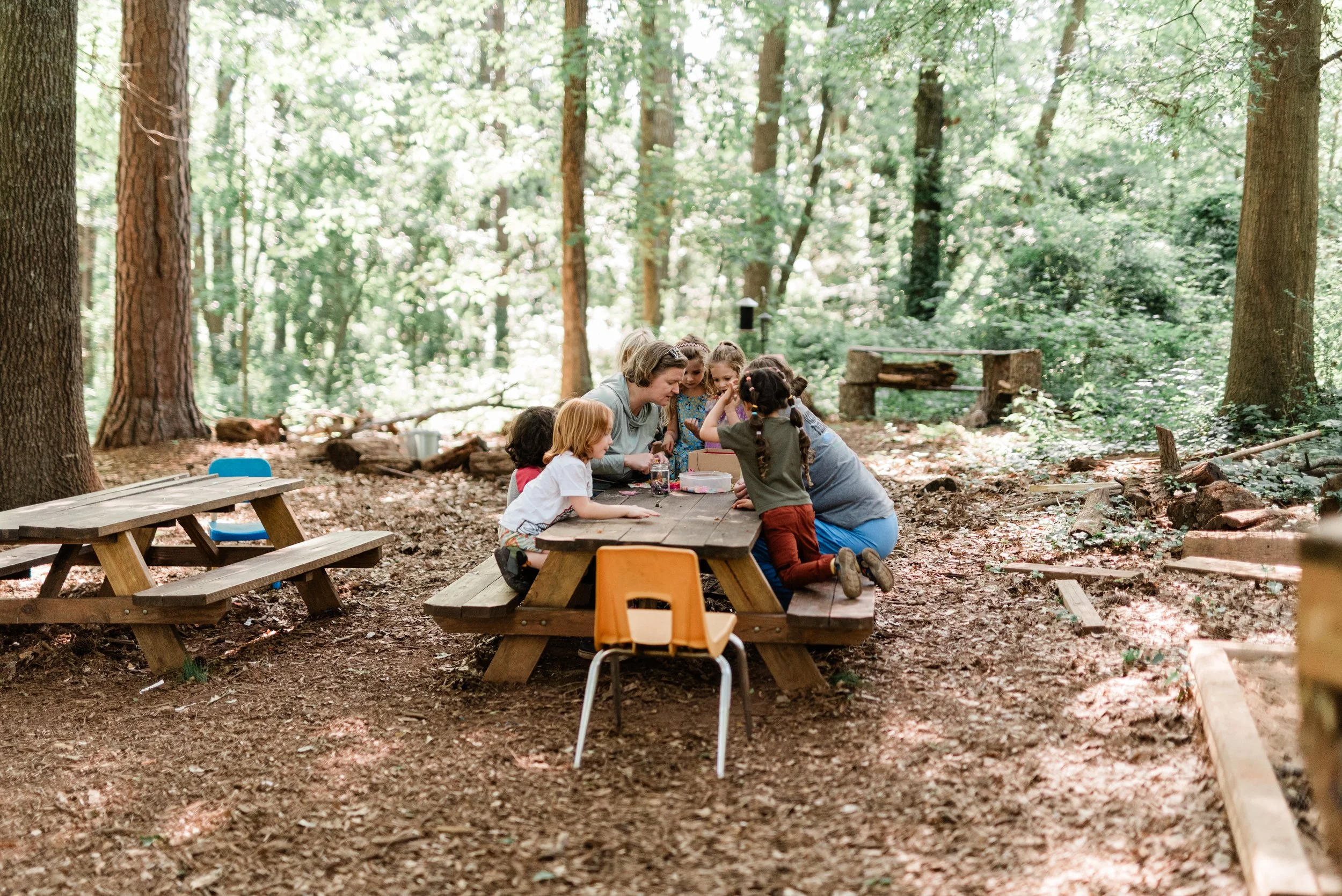 A group of children and two adults sitting at a picnic table in a forest school.