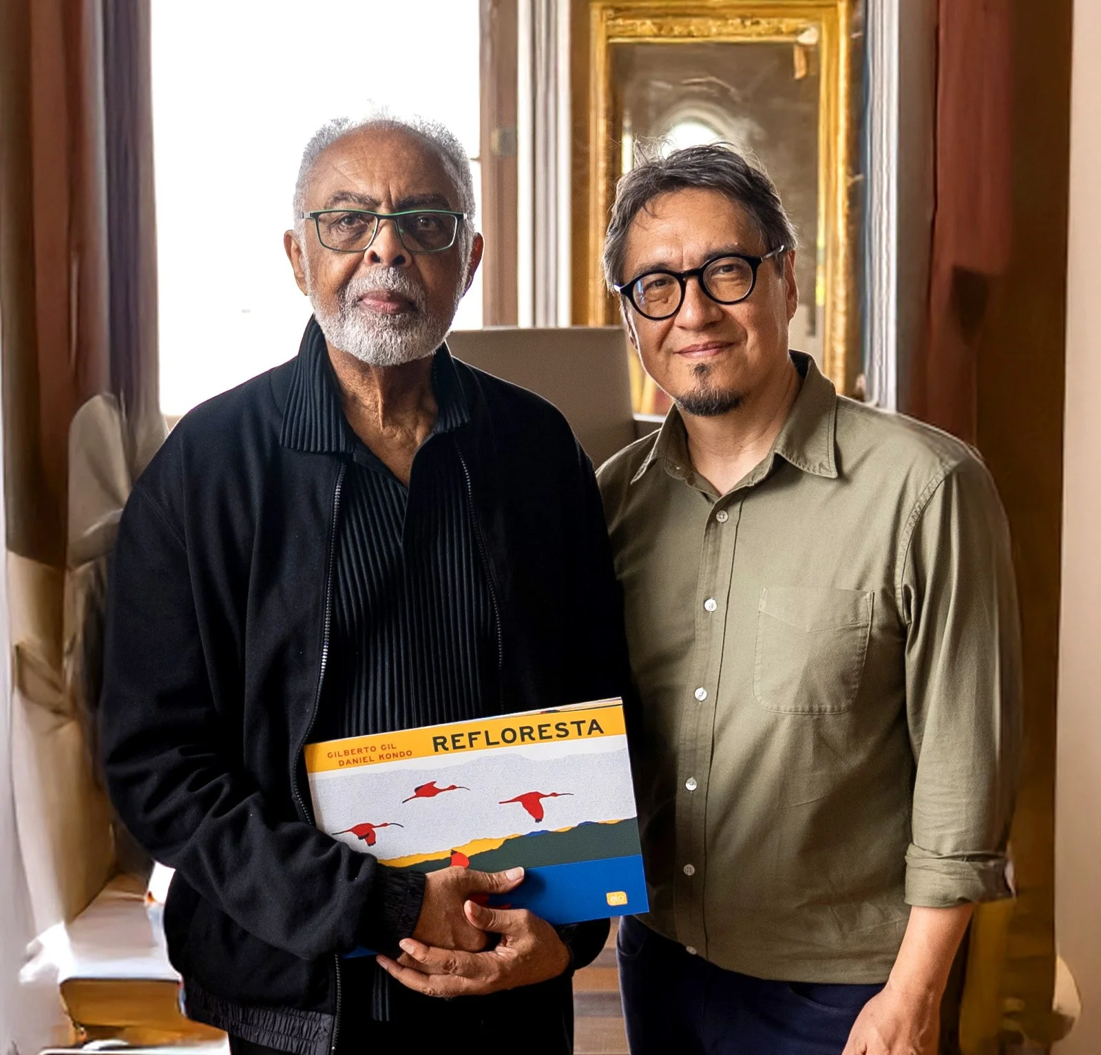 Daniel Kondo e gilberto Gil. Gil segura o livro em suas mãos. está de casaco preto. Daniel Kondo, à direita, esta de camisa verde. ambos de pé, com a janela interna do Theatro da Paz ao fundo