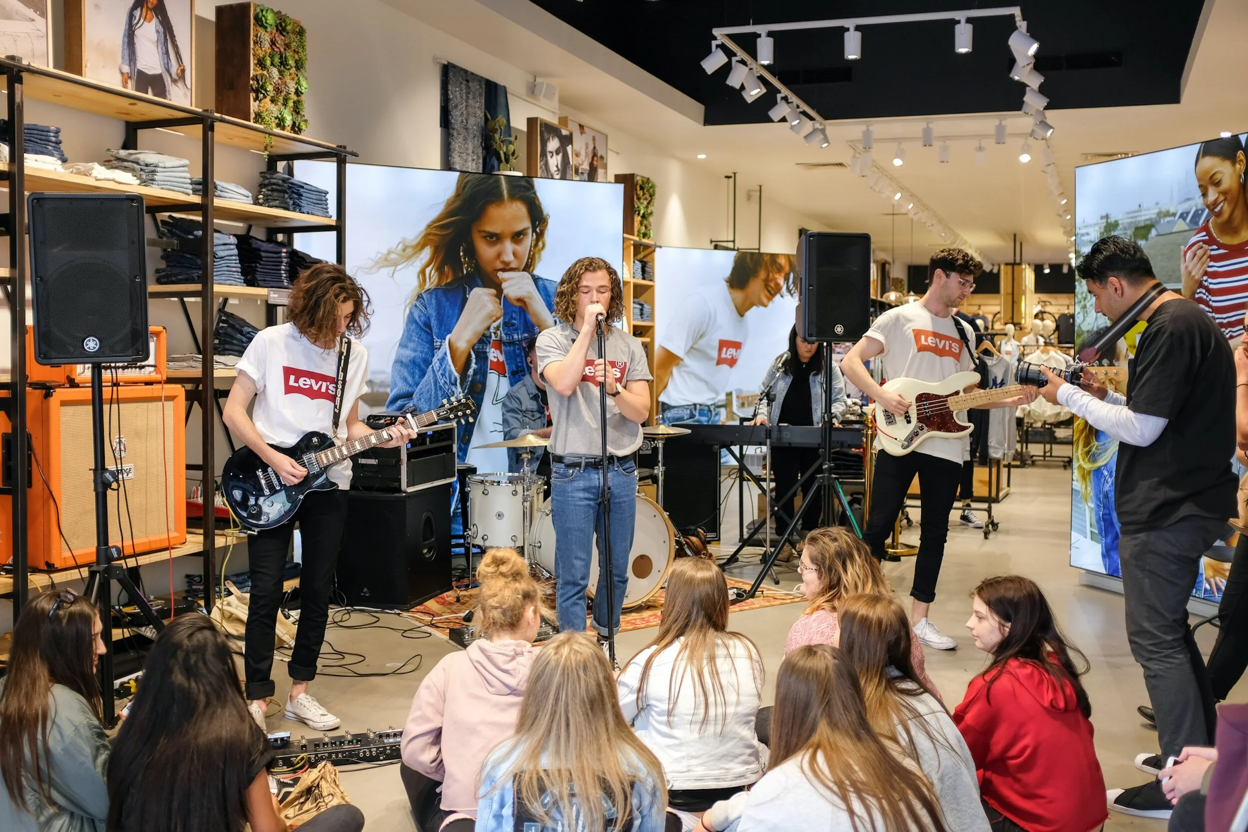 A band performs on an indoor stage in a retail store, with five members playing various instruments and singing, while an audience of young women watches seated on the floor. The background features large digital screens and shelves of clothing.