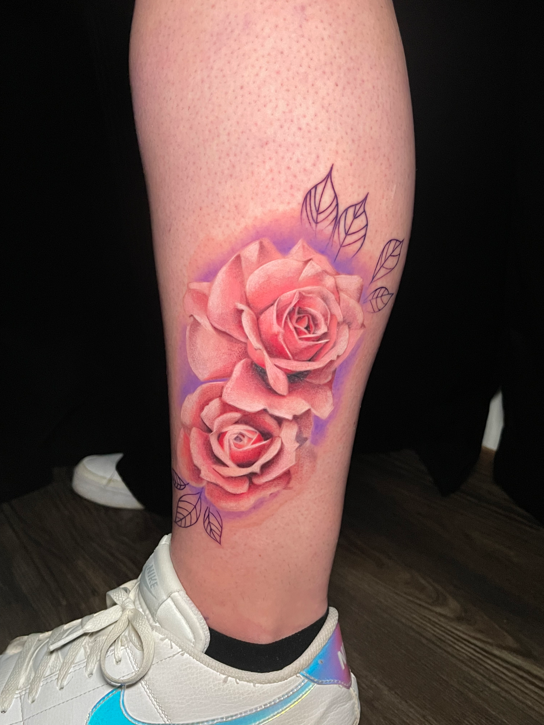 Tattoo of two pink roses on a leg with violet shading and outline of leaves, near a white sneaker.