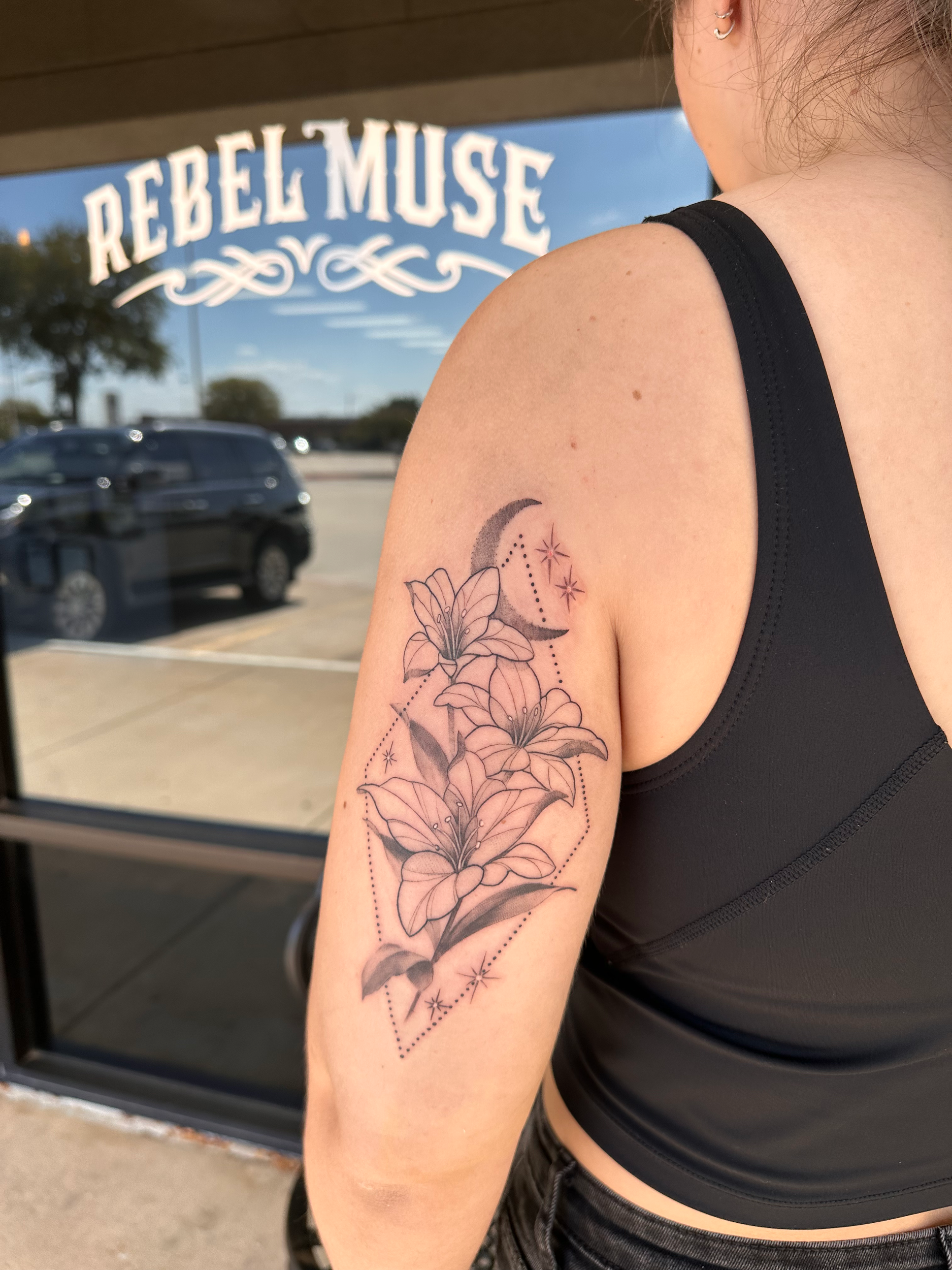 Person with floral tattoo featuring lilies and crescent moon design on upper arm outside tattoo shop.