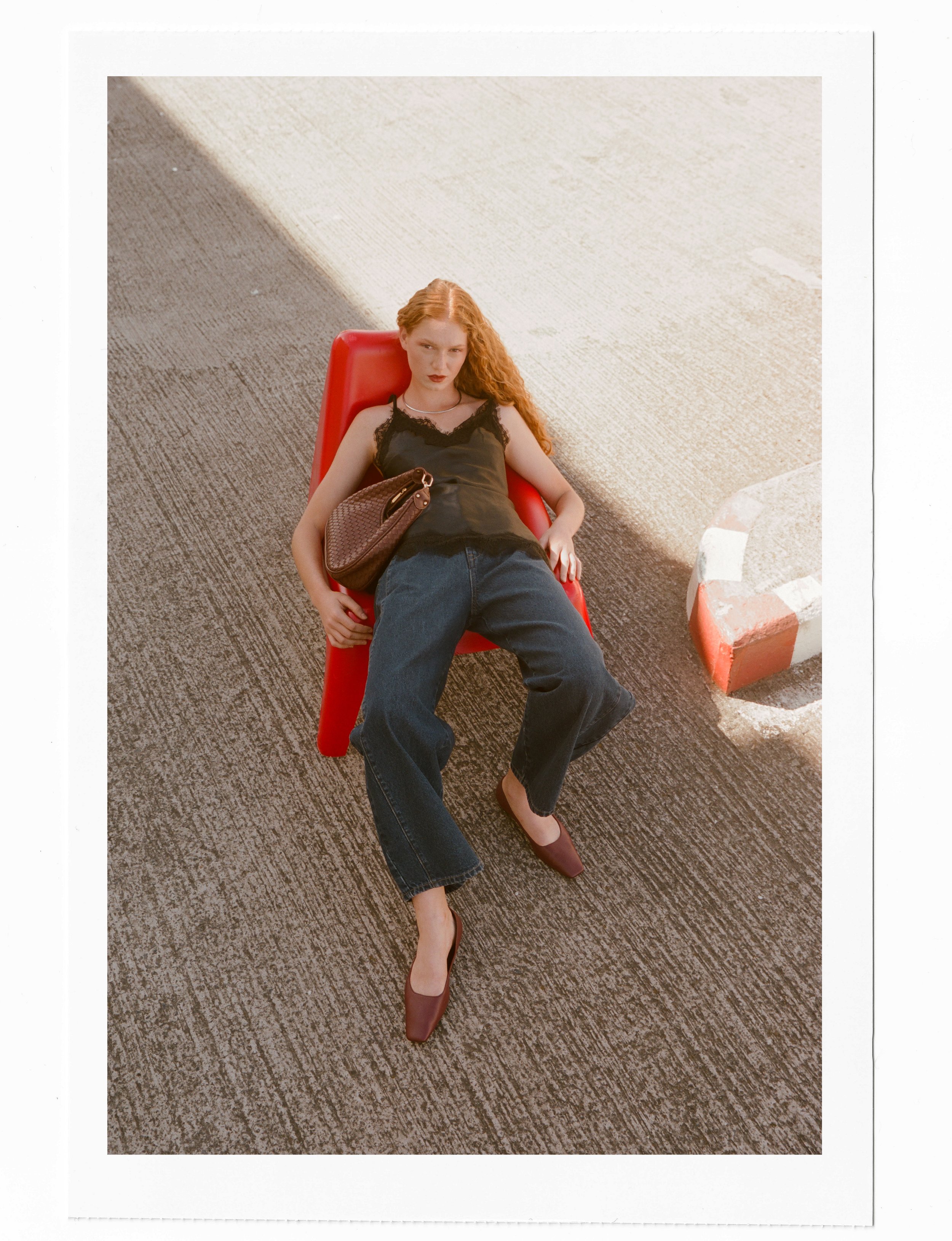 A woman with long curly red hair lying on a red lounge chair outdoors, with one leg crossed over the other, wearing a black lace top, dark jeans, and brown pointed shoes, holding a purse on her lap, with a textured outdoor surface and a small brick border nearby.