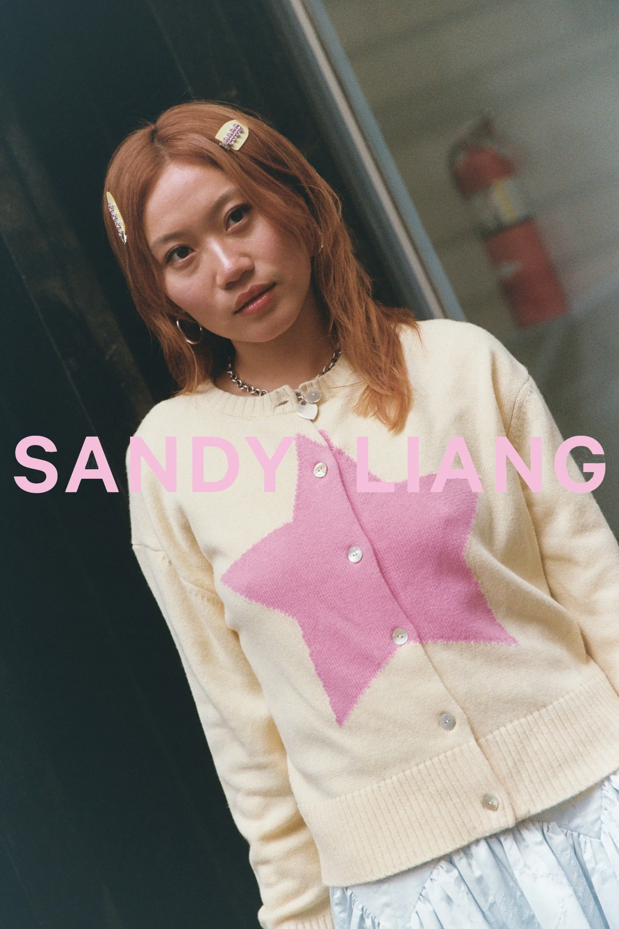 Close-up of a young woman with light brown hair wearing hair clips, a chain necklace, and a cream-colored cardigan with a pink star pattern, standing in front of a black door with a fire extinguisher in the background.