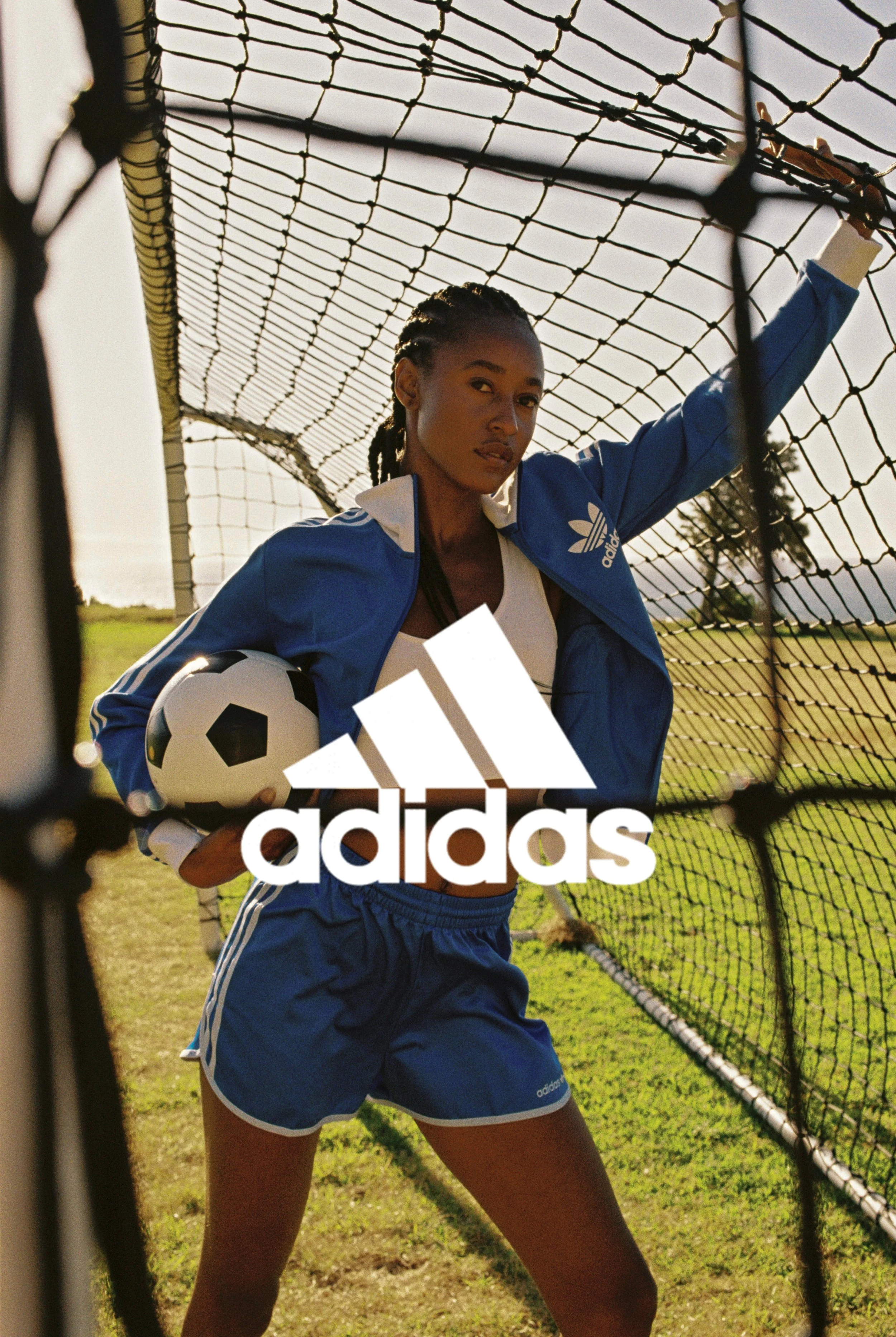 A woman in blue Adidas sportswear posing on a soccer field, holding a soccer ball, and standing in front of a soccer goal net under a partly cloudy sky.