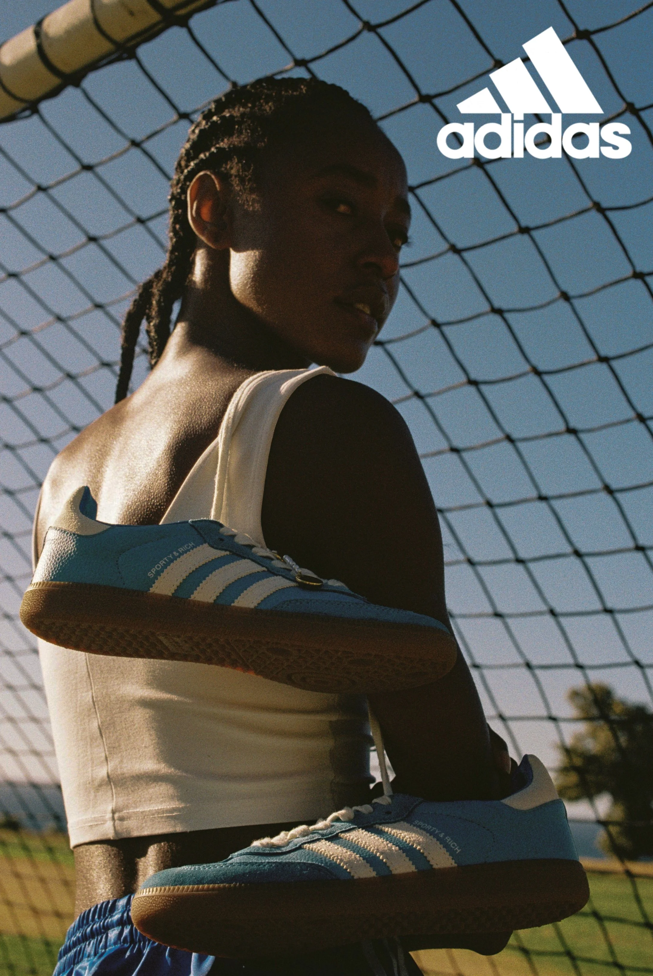 Close-up of a person with braided hair wearing Adidas sneakers and a white tank top, holding a basketball on a sports court with a fence and clear blue sky in the background.