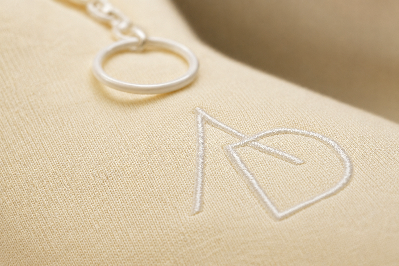 Close-up of an embroidered logo on a beige fabric, featuring a stylized letter 'R' and a circular ring attached to a chain. zipper, quarter zip ;luxury brand, street wear