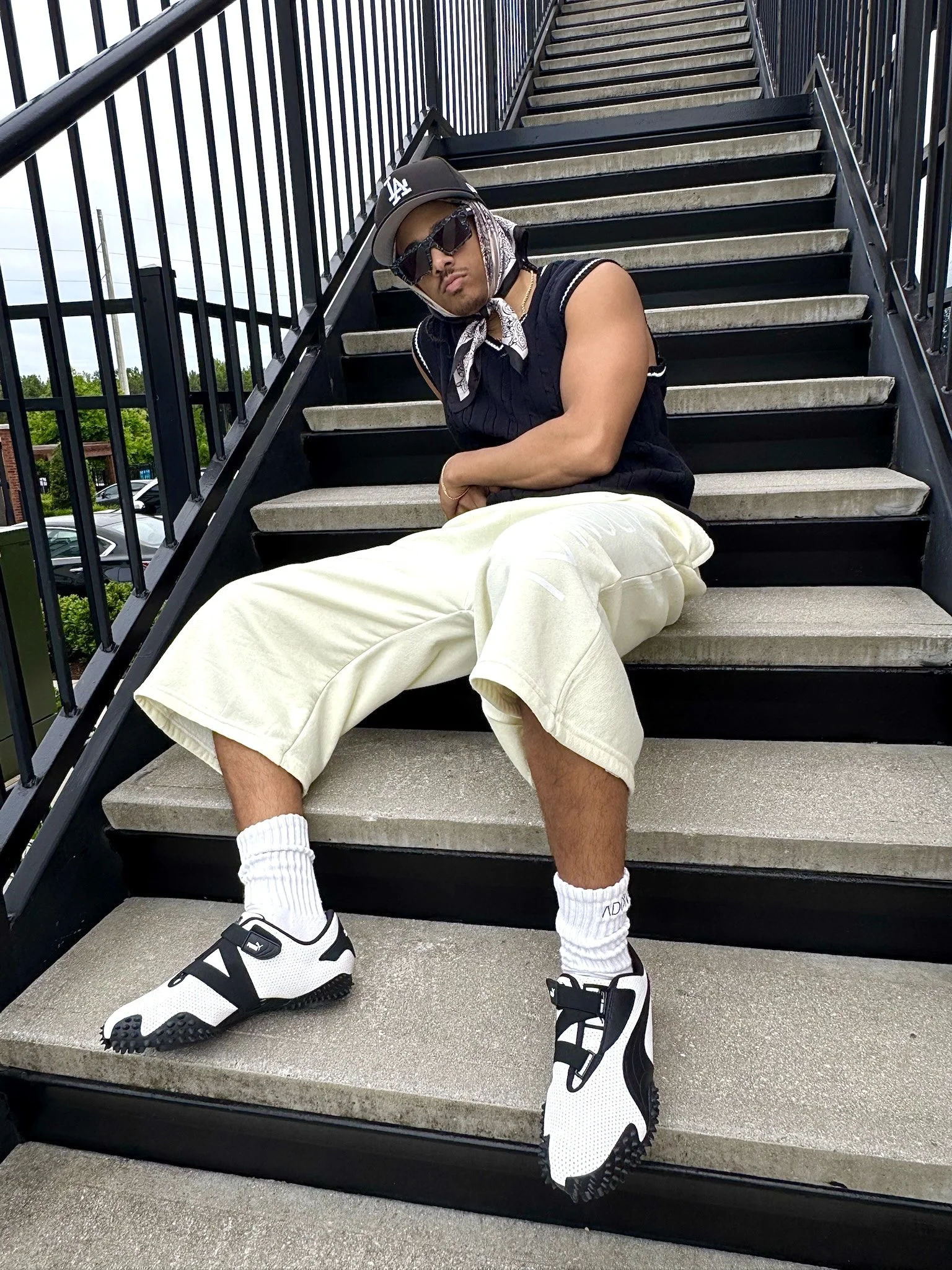 A man sitting on outdoor staircase, dressed in a black sleeveless shirt, light beige shorts, white athletic shoes with black accents, black sunglasses, a black baseball cap with white lettering, a patterned scarf around his neck, and a gold chain. He