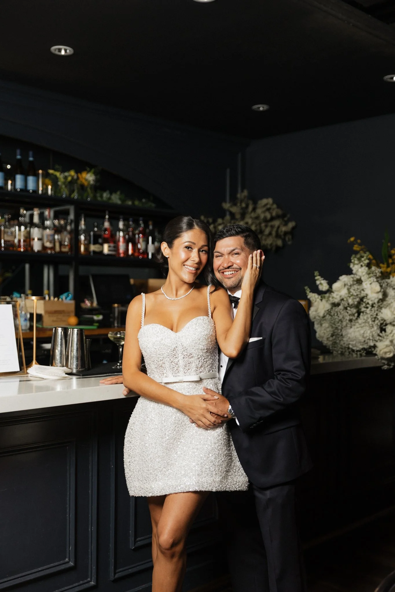 A happy bride and groom stand together at a bar, smiling and embracing during their wedding celebration, with flowers and bottles of alcohol in the background.