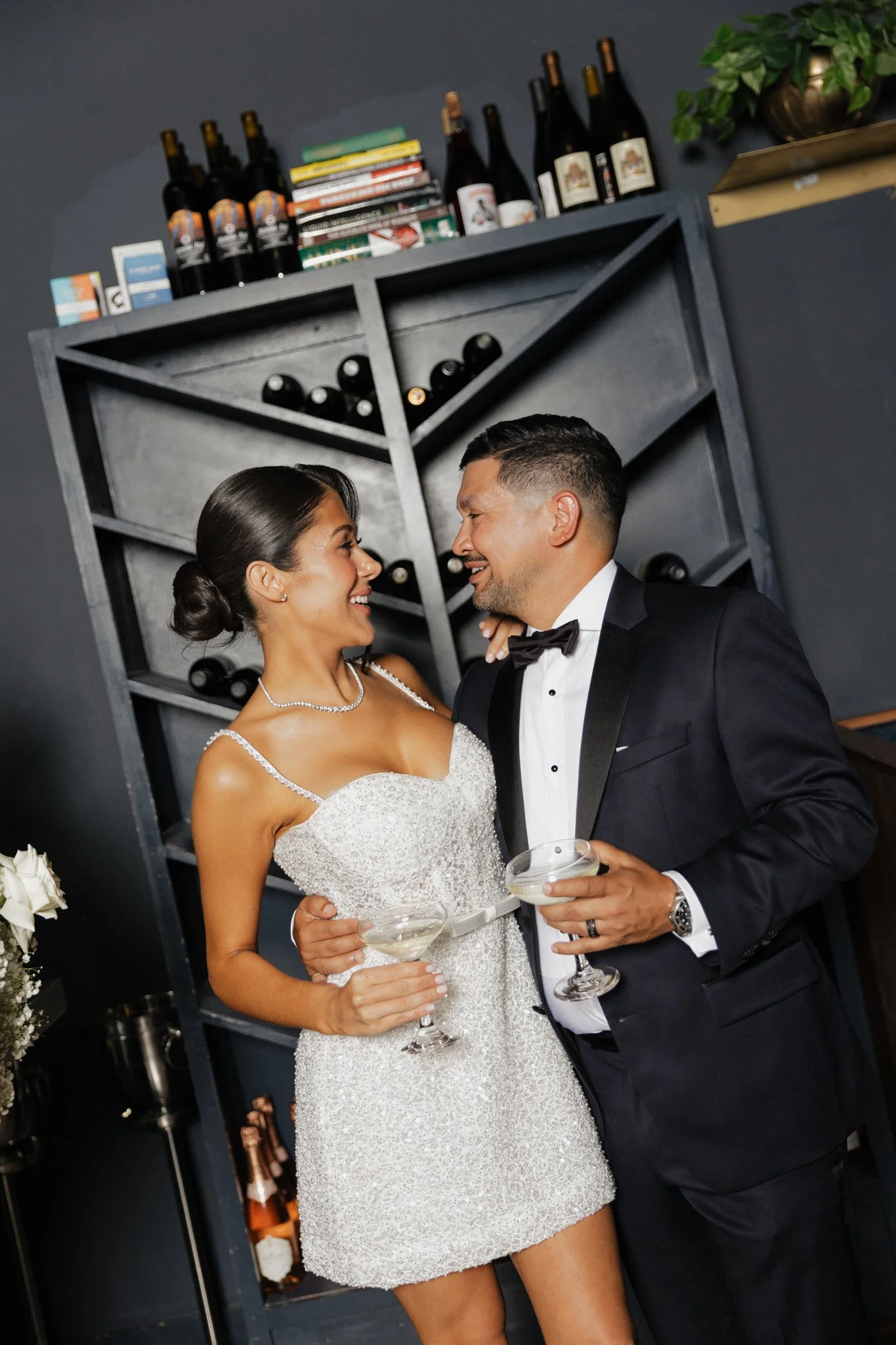 A bride and groom in formal attire dancing and holding champagne glasses at their wedding reception, with a wine rack and books in the background.