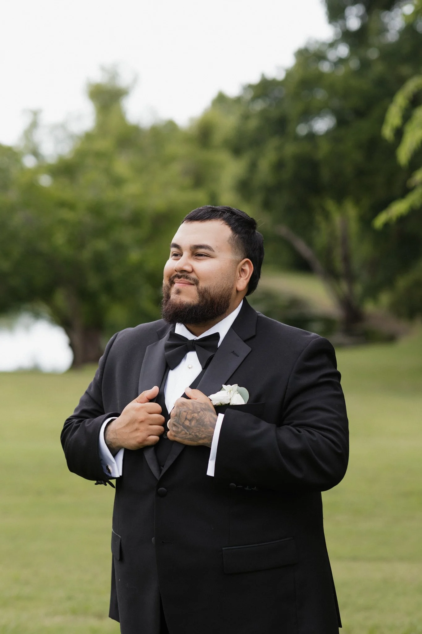 Groom in a black tuxedo with a bow tie, standing outdoors with a green landscape and trees in the background, adjusting his jacket.