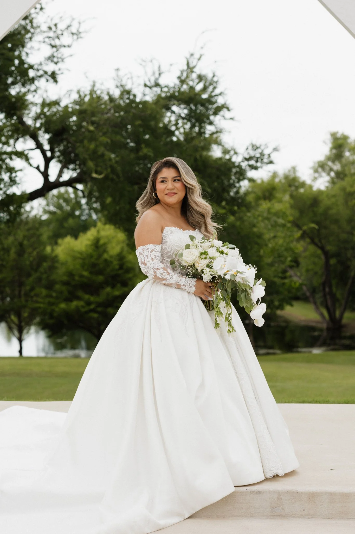 A bride in a white wedding gown holding a bouquet of white flowers outdoors, with green trees and a pond in the background.