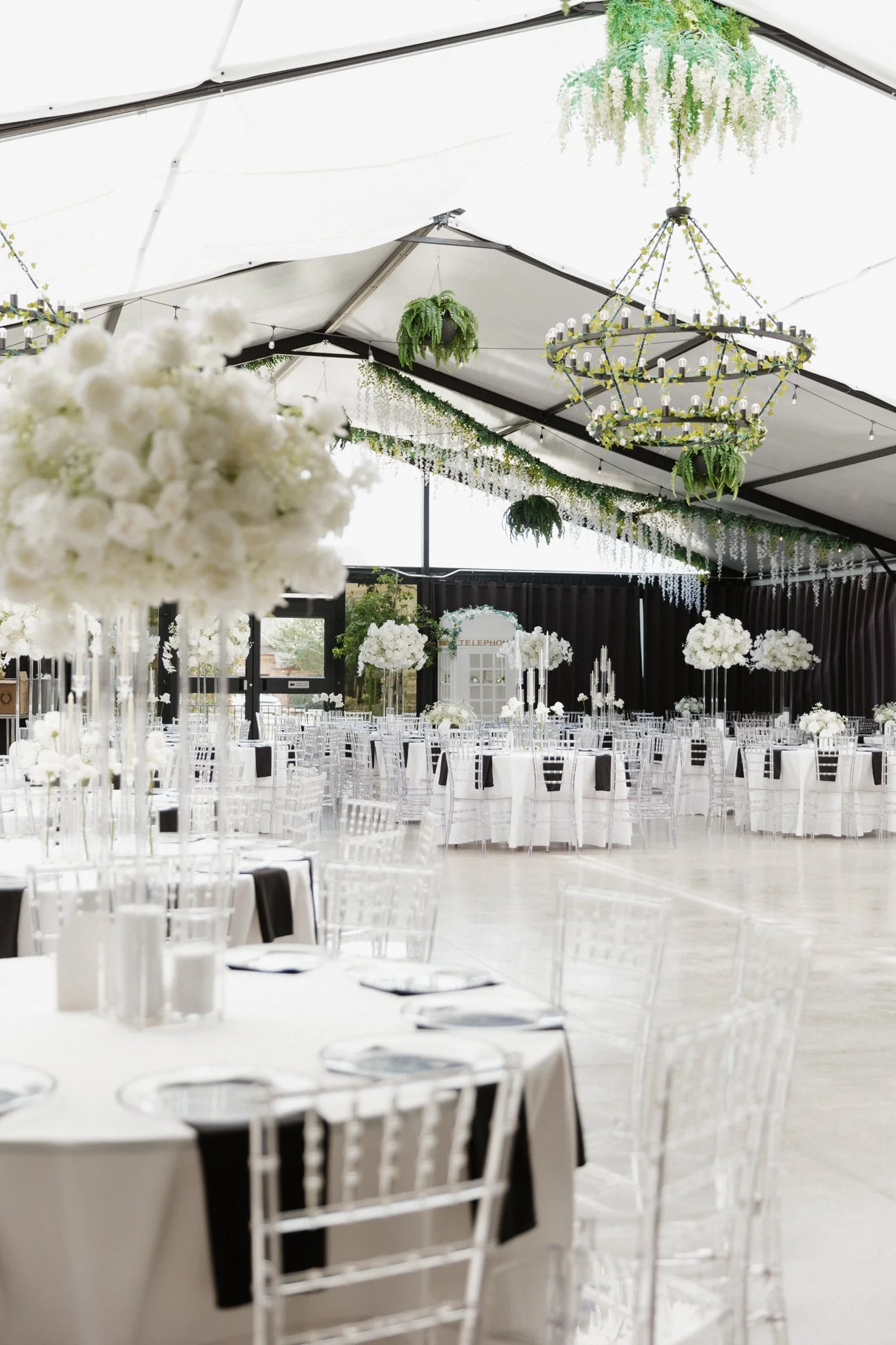Elegant event space decorated with white floral arrangements, hanging greenery, chandeliers, and black and white table settings for a formal celebration.