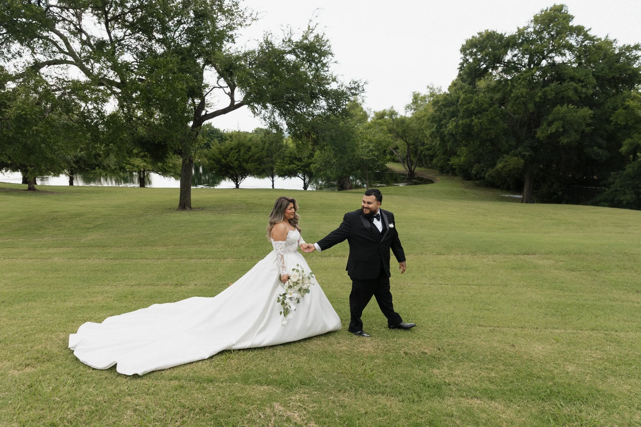 A bride and groom holding hands on a grassy field near a lake, surrounded by trees, during a wedding photoshoot.