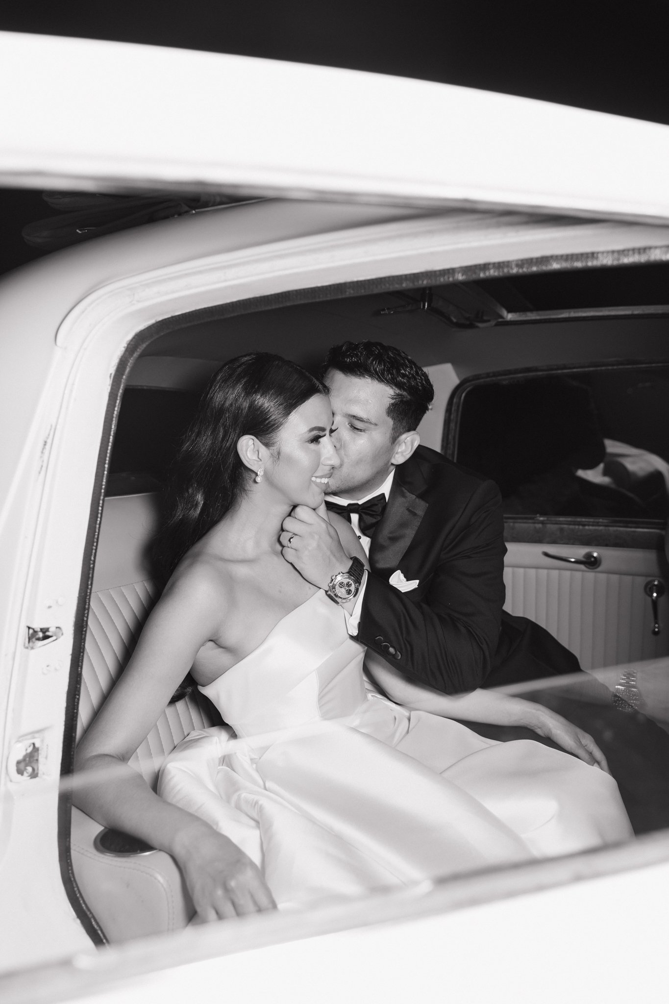 A bride and groom sitting inside a vintage car, sharing a tender moment. The groom is kissing the bride on her forehead, and they are both smiling. The bride is wearing earrings and a strapless dress, while the groom is dressed in a tuxedo.