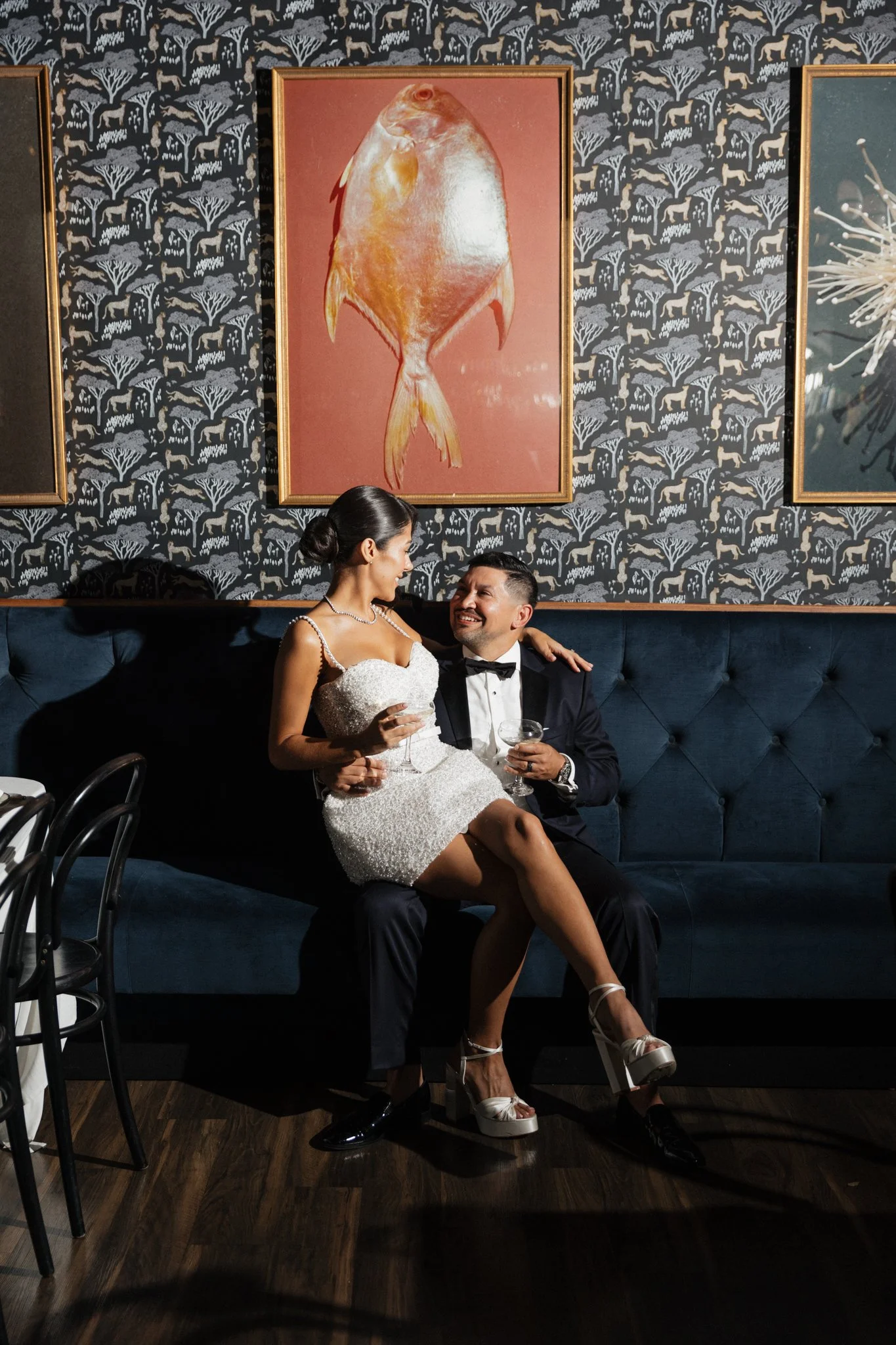A man and woman dressed in formal attire sitting on a dark blue velvet couch, looking at each other and smiling. The woman is wearing a white lace dress and high heels, sitting on the man's lap. The man is wearing a black tuxedo with a bow tie. They 