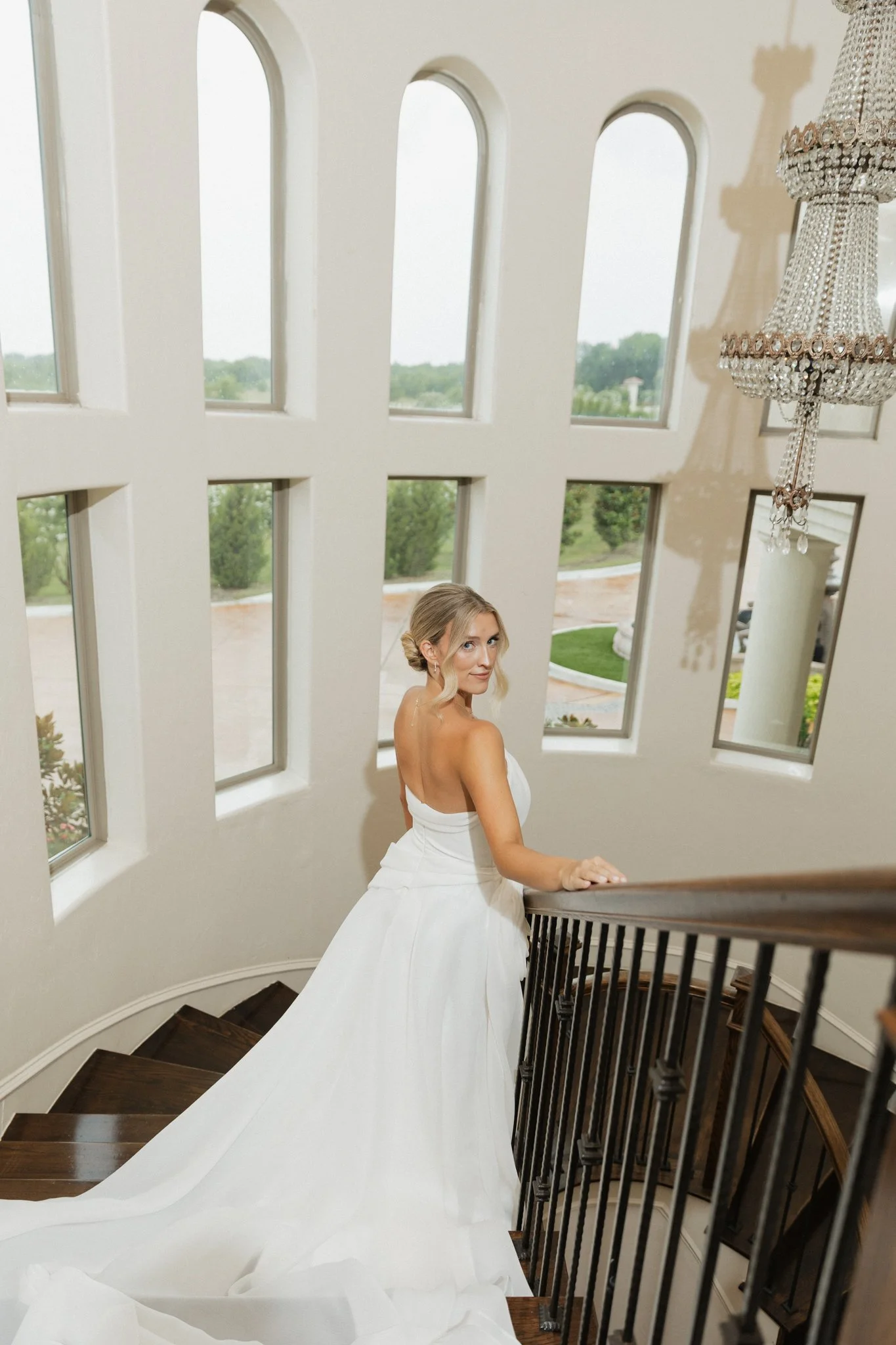 A bride with blonde hair in an updo, wearing a strapless white wedding gown, standing on a staircase inside a building with large arched windows and a crystal chandelier.
