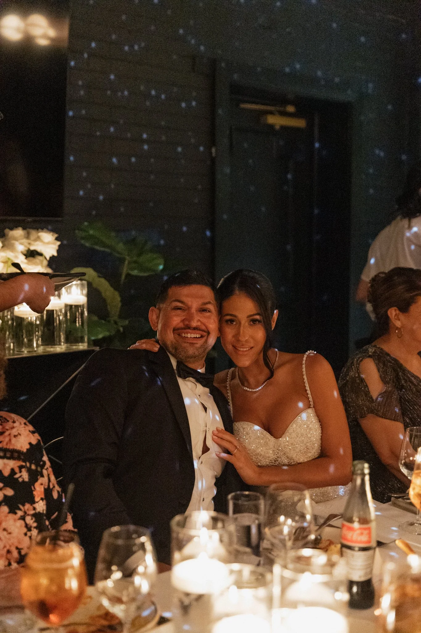 A couple dressed in formal attire, with the man in a tuxedo and the woman in a wedding dress, smiling and posing at an elegant dinner event.
