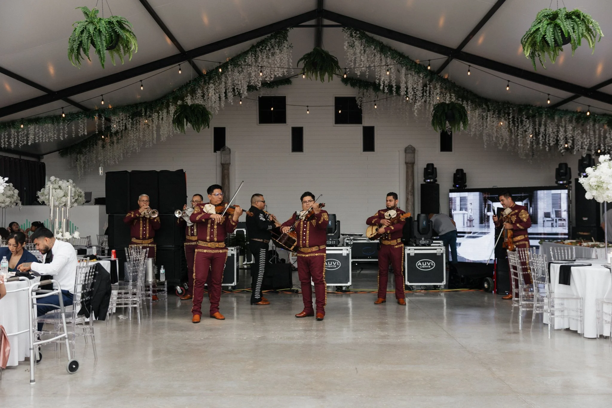 Band performing at a celebration inside a decorated event hall with hanging greenery and string lights.