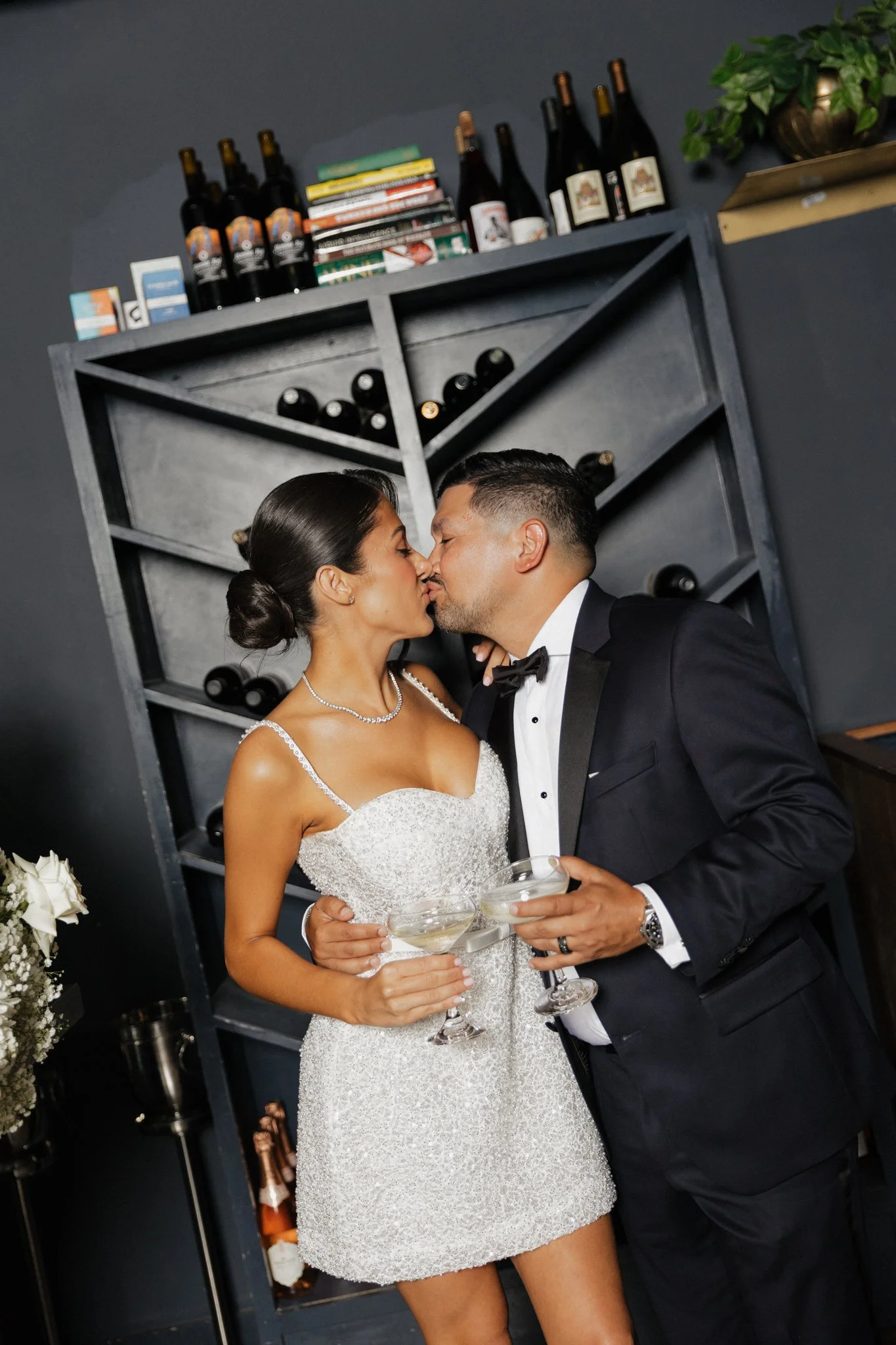 A bride and groom are about to kiss at a wedding reception, holding glasses of champagne, standing in front of a gray wall with shelves of wine and books.