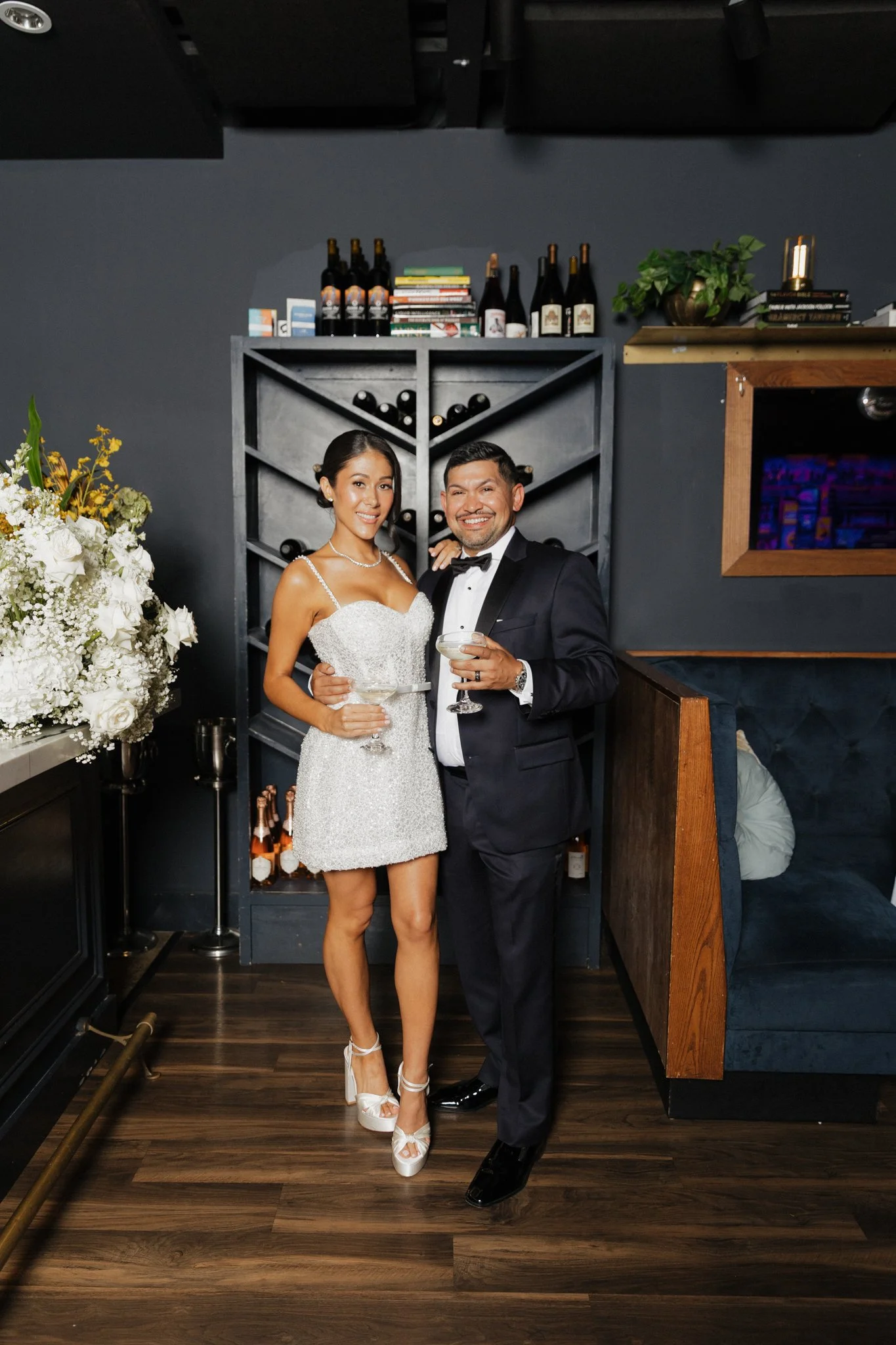 A bride and groom in wedding attire standing together indoors, holding cocktail glasses, smiling at the camera, with a dark wall, shelves with wine bottles, and floral arrangements in the background.