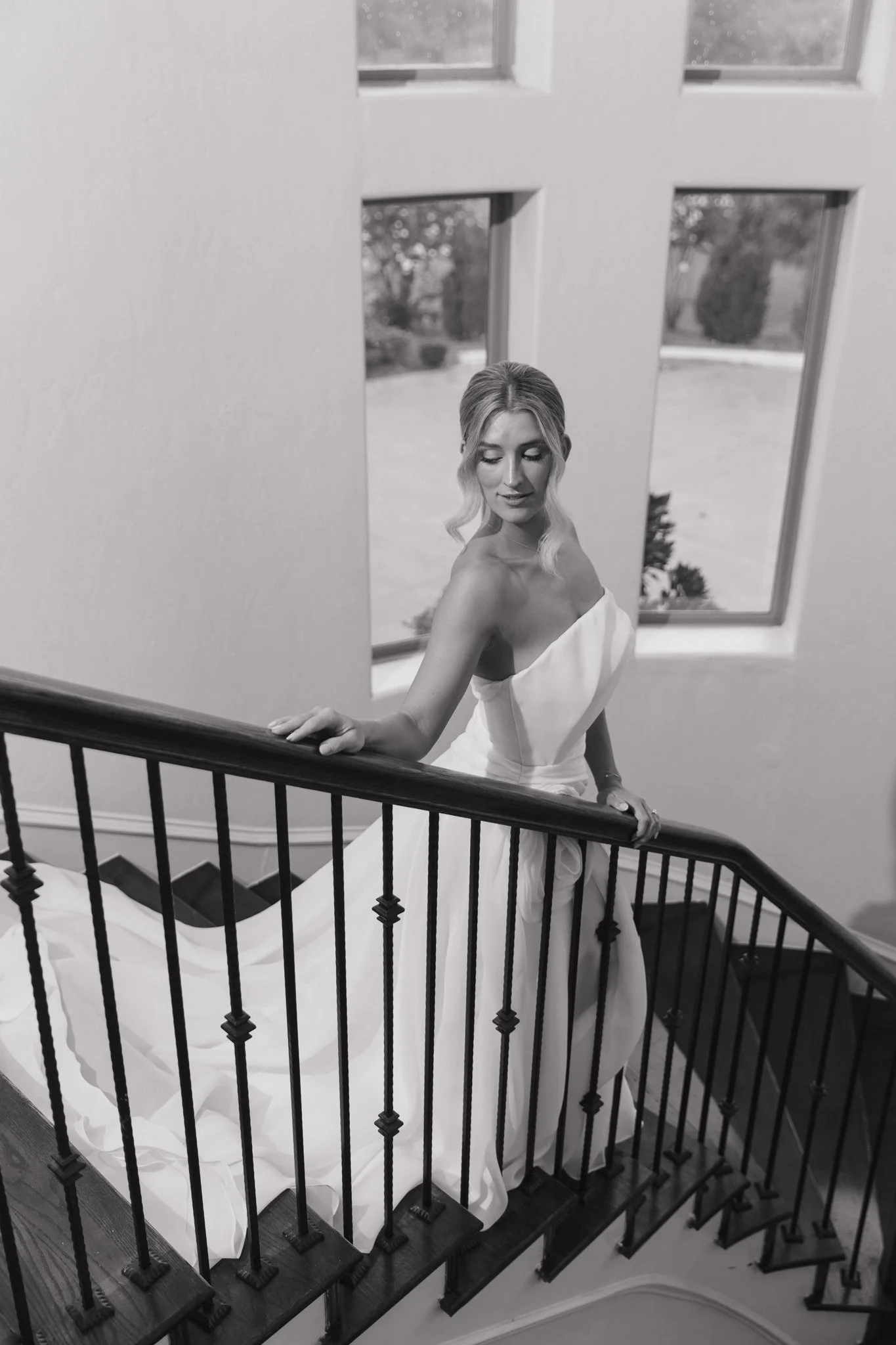 Black and white photo of a woman in a strapless wedding gown standing on stairs near a window, holding the railing with her left hand.