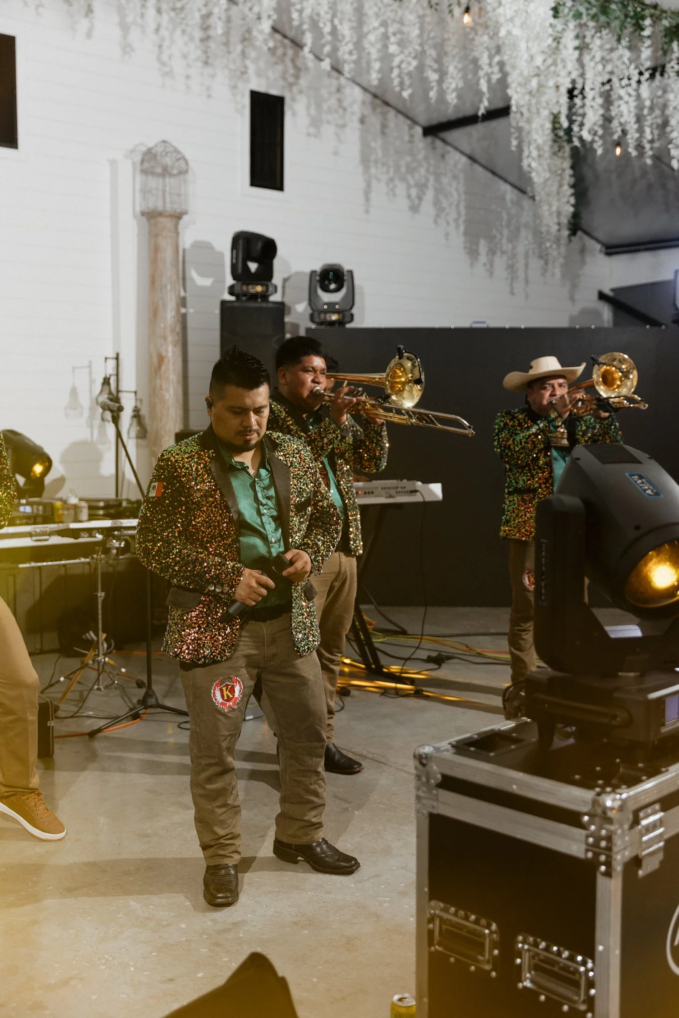 Musicians in sequined jackets playing trombones during a live performance indoors, with lighting and sound equipment visible.