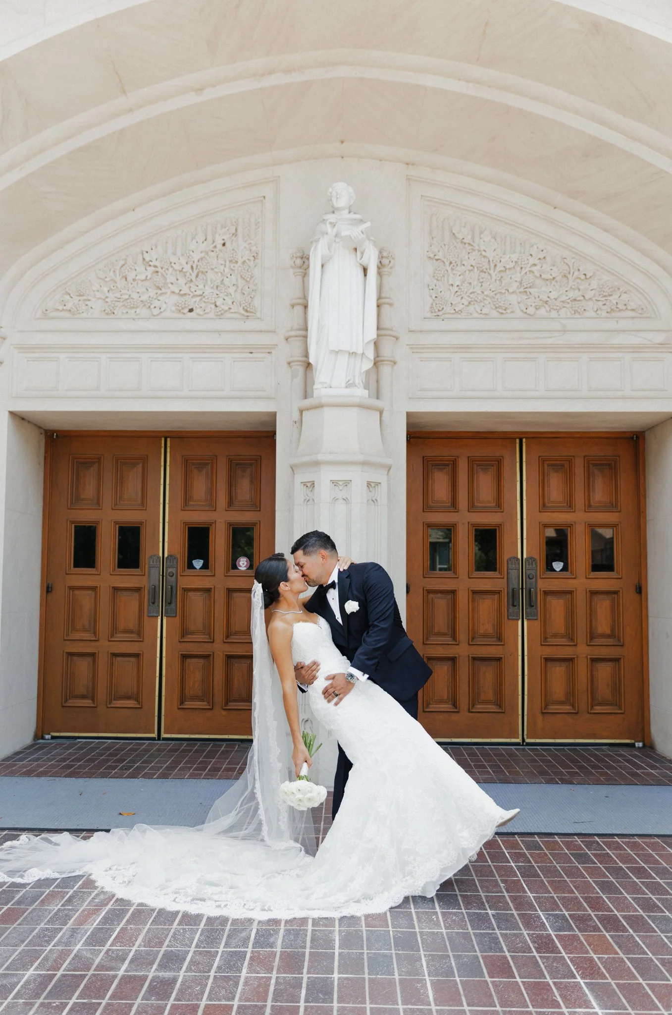 A newlywed couple sharing a kiss in front of a church entrance with large wooden doors and ornate architectural details, including a white statue above the doors. The bride is in a white wedding gown and holding a bouquet, and the groom is in a black